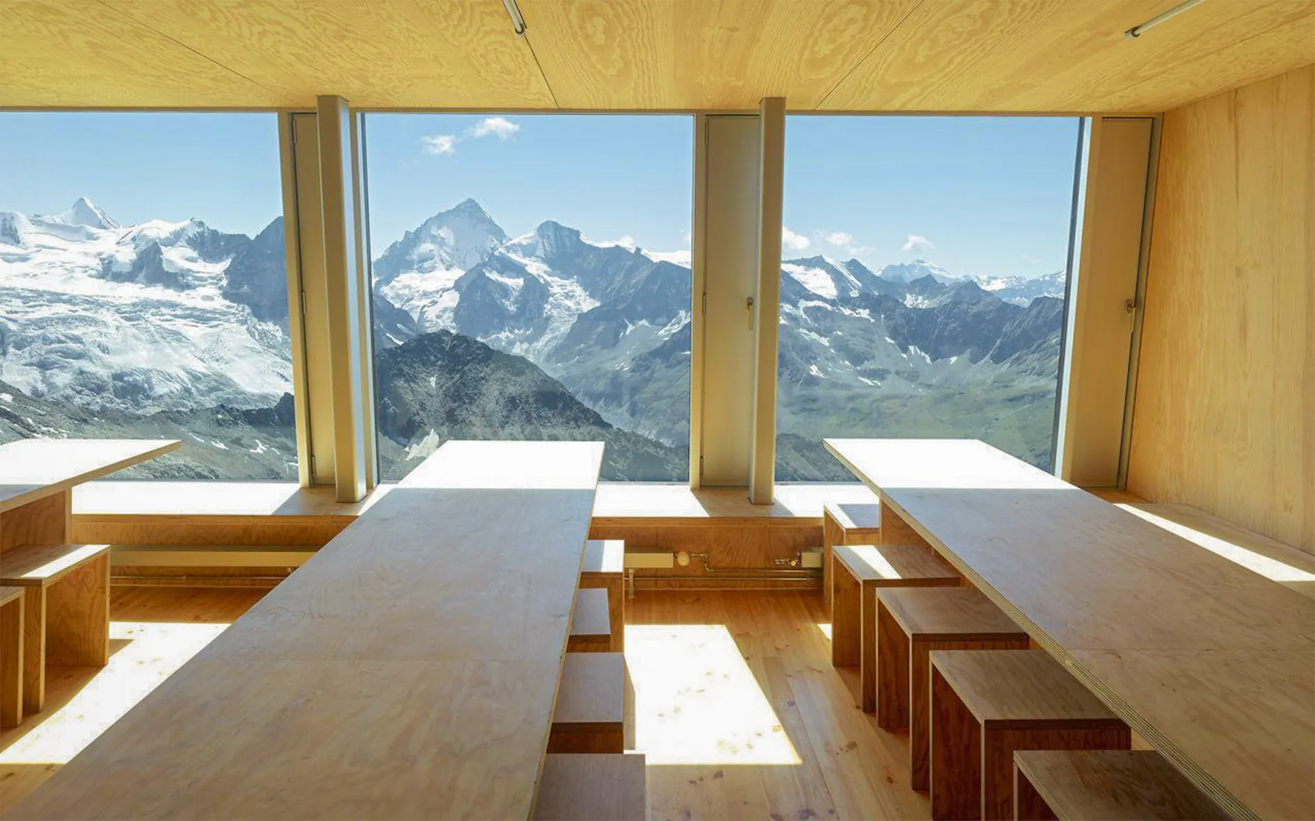A minimalist interior with long light wood tables and matching benches. Large windows offer panoramic views of majestic mountains, partly covered in snow and surrounded by greenery.