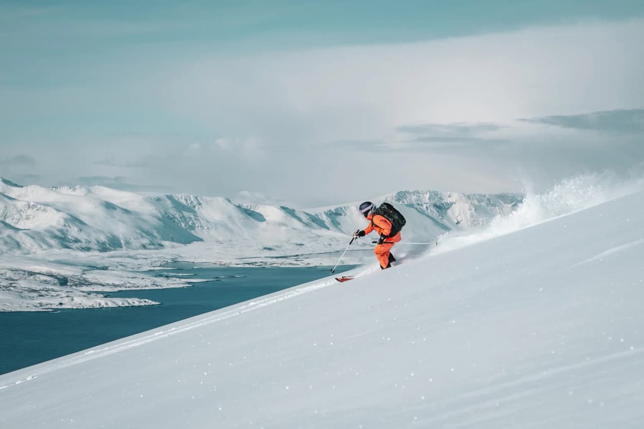 A skier descends a snowy slope, approaching a spectacular mountain landscape. He is wearing a bright orange wetsuit and a rucksack, and his skis are tracing a line in the fresh snow. In the background, mountains