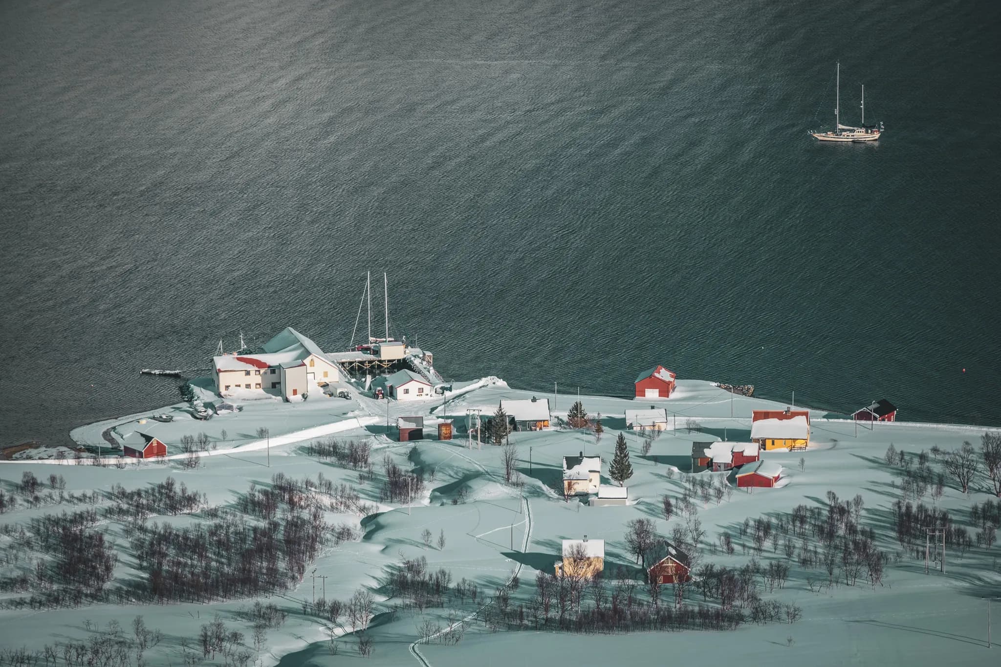Un paysage hivernal surplombant une petite colonie située au bord de l'eau. Des maisons aux toits colorés, principalement rouges et jaunes, sont dispersées sur des collines recouvertes de neige. Un quai et des
