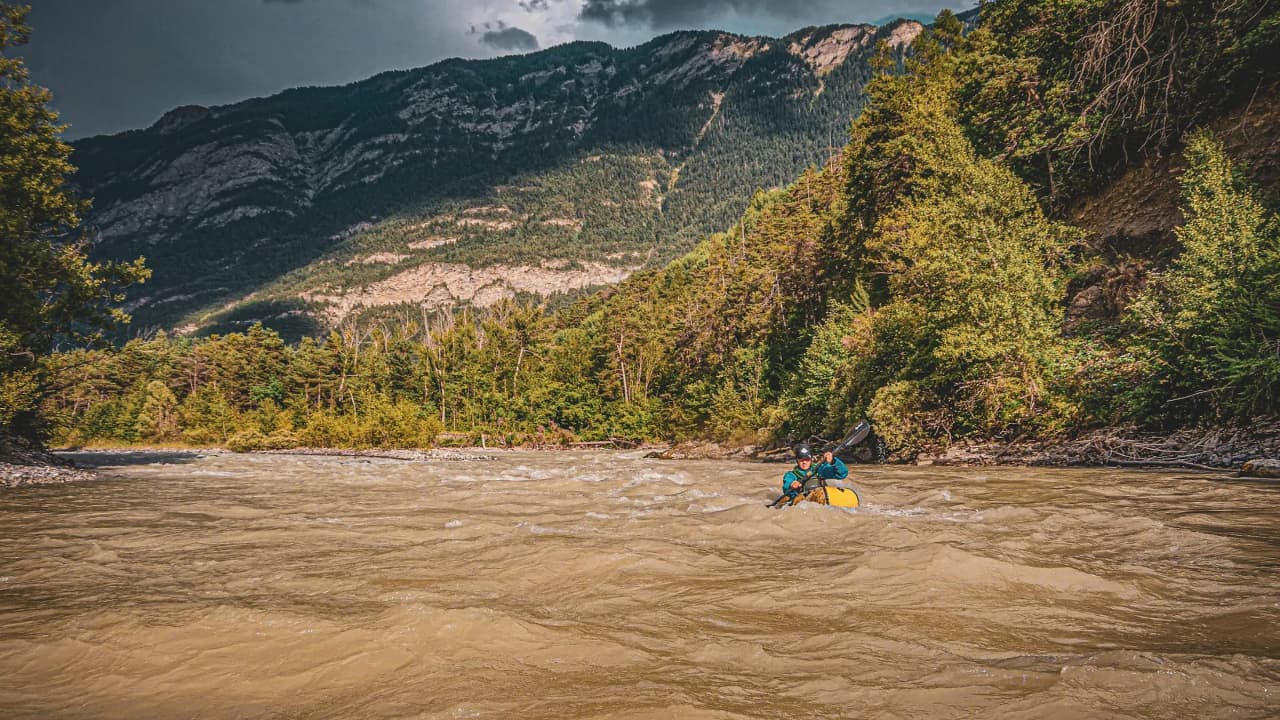 Een avonturier in een packraft vaart een rivier af die wordt omringd door groene bergen, onder een indrukwekkende hemel.