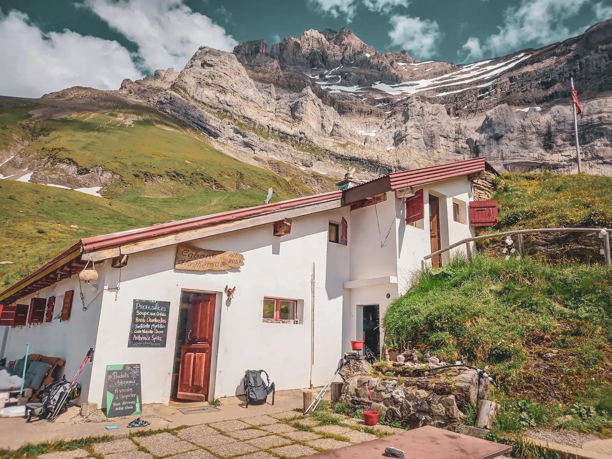 A mountain chalet set at the foot of towering rocky cliffs, with snow-capped peaks in the background. The building is white with a red roof and wooden shutters. A wooden sign