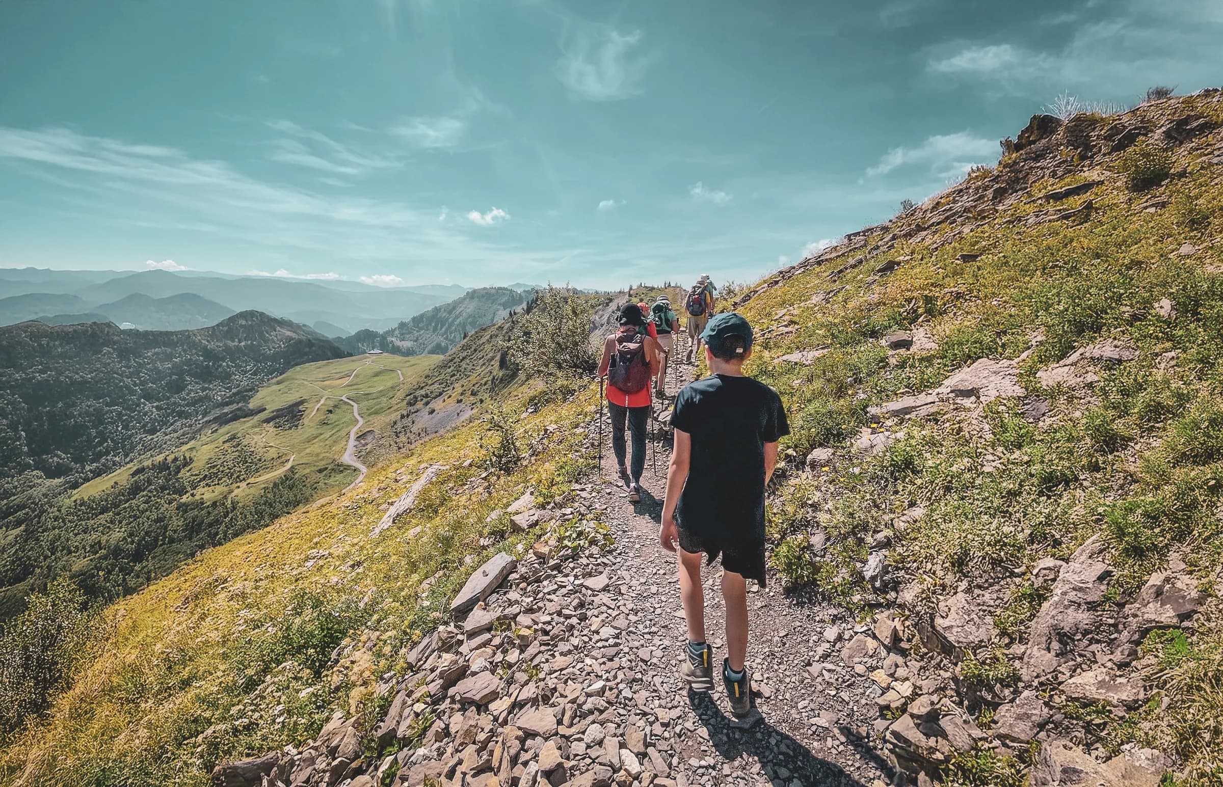 A group of people walk along a narrow mountain path, surrounded by lush green landscapes and hills. The path is rocky, and panoramic views of the mountain ranges in the distance stretch as far as the