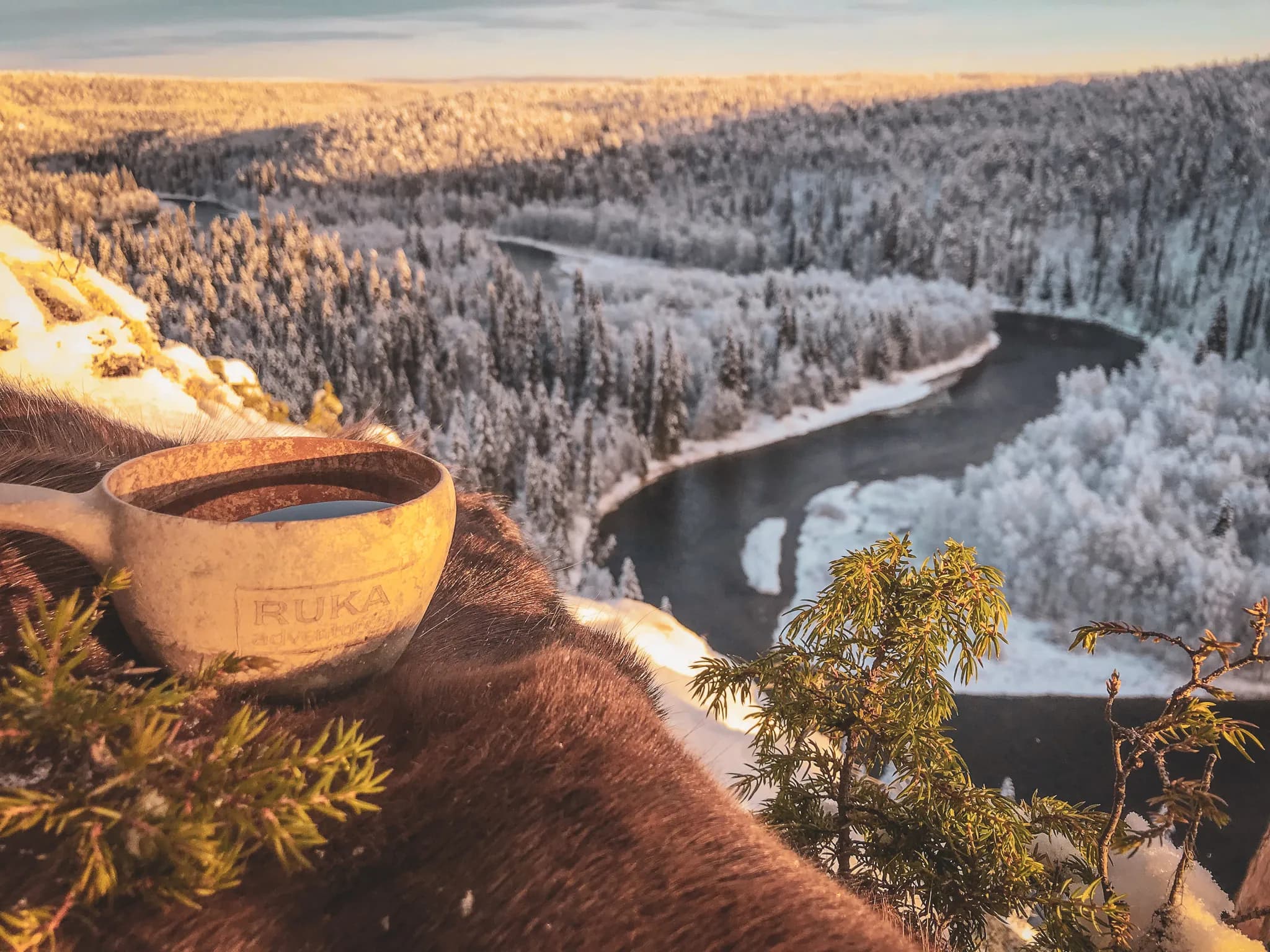 A spectacular winter landscape offers a view of a wooded valley covered in snow. In the foreground, a ceramic mug bearing the "RUKA" logo, probably containing a hot drink, sits on a blanket of snow.