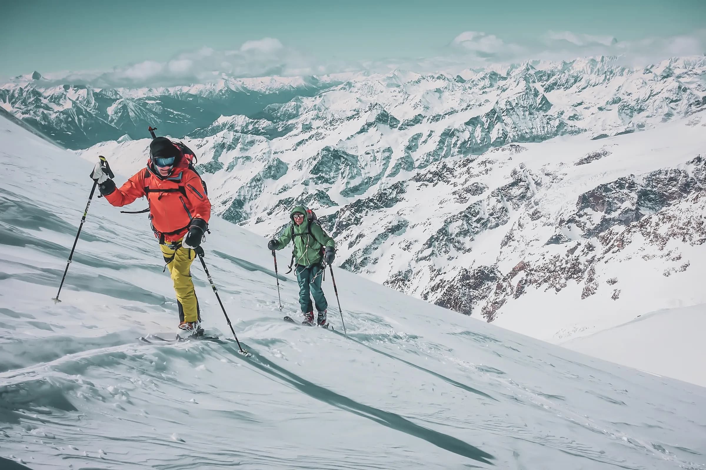 Two skiers navigate a snowy slope, surrounded by majestic mountains. The skier on the left is wearing a bright orange jacket and bright yellow trousers, while the skier on the right is dressed in a bright yellow jacket.