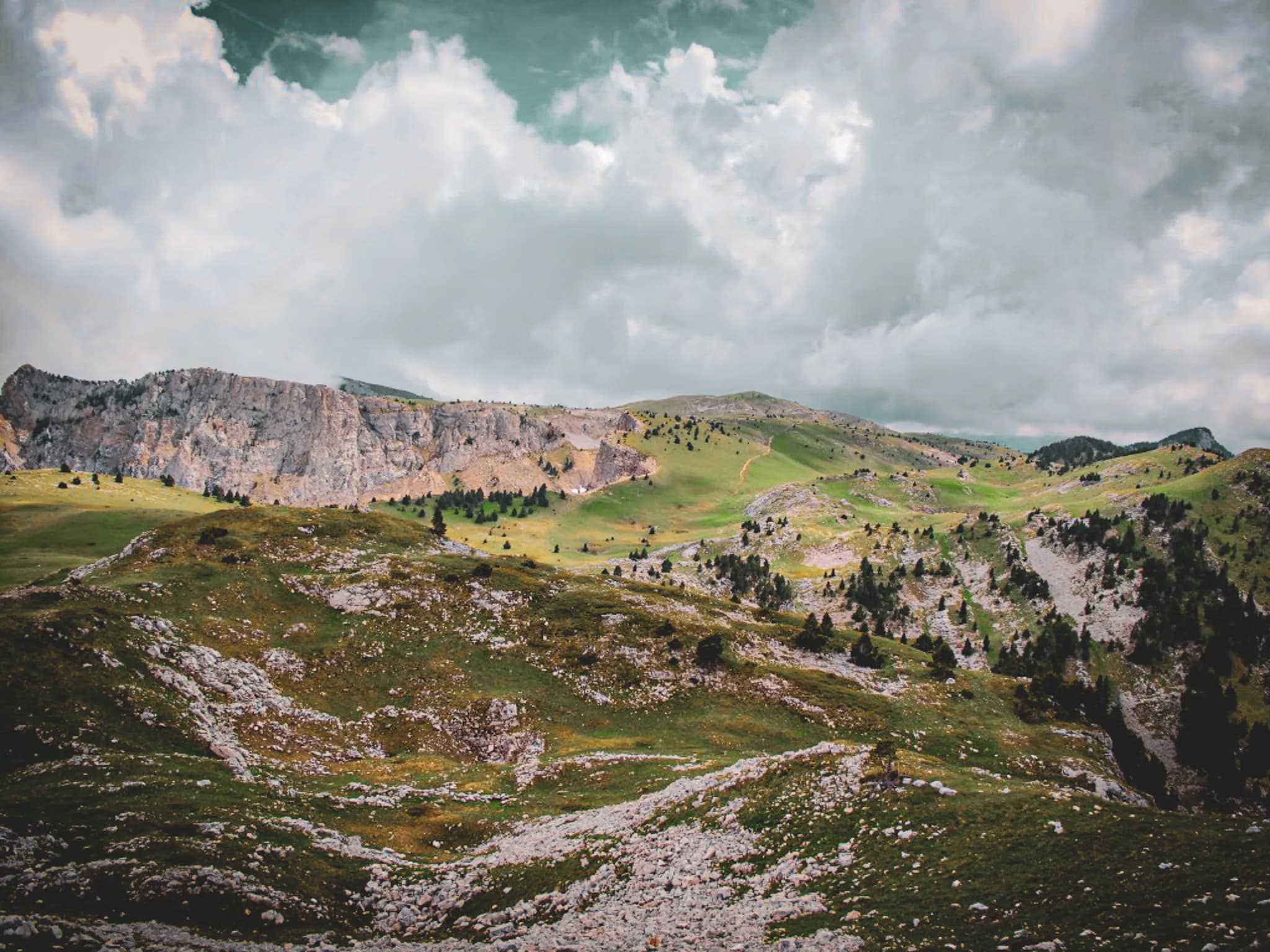 A mountainous landscape with green hills and scattered rocks. In the background, sheer cliffs dominate the panorama, while a cloudy sky with shades of grey and blue adds to the beauty of the scenery.