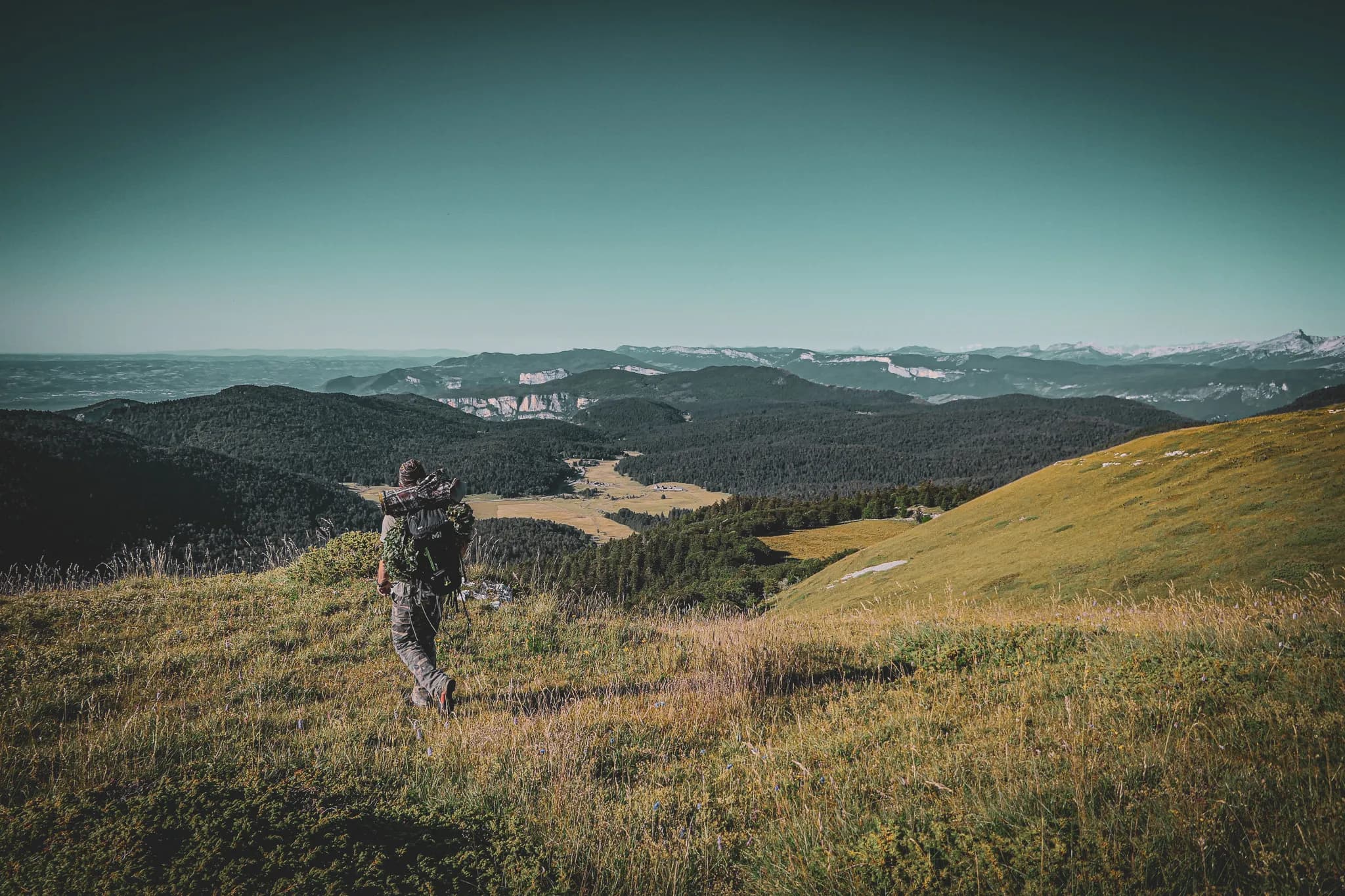 A man is walking along a green path overlooking a vast valley and distant mountains. He is carrying a rucksack, loaded with equipment, and stares into the distance, captivated by the spectacular landscape. The scene is bathing