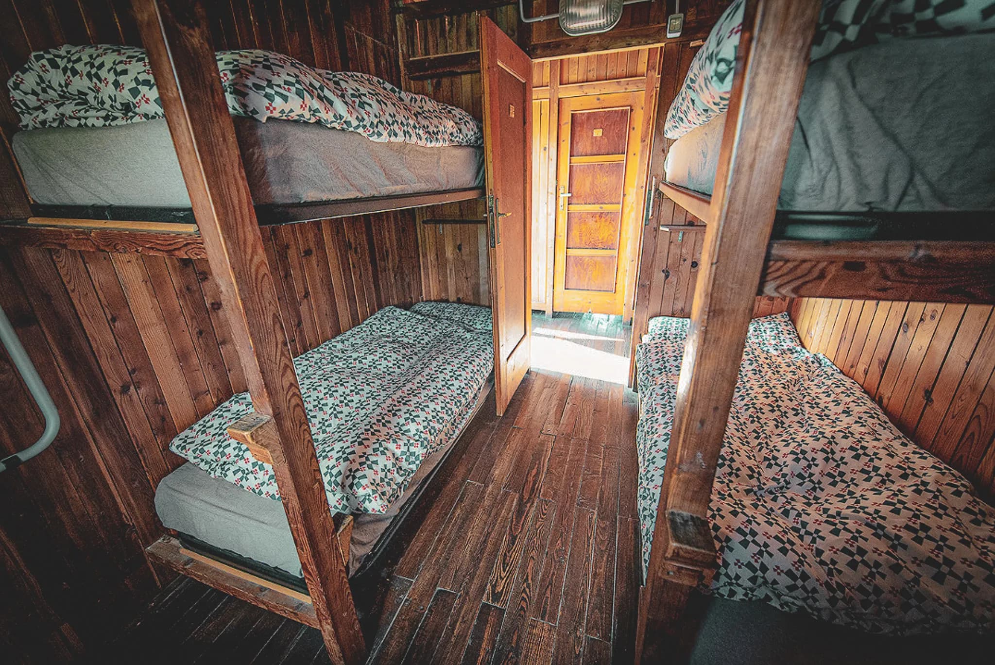 A wooden room with bunk beds and mattresses covered with sheets decorated with floral motifs. The decor is rustic, with dark wood walls and a wooden floor. Two