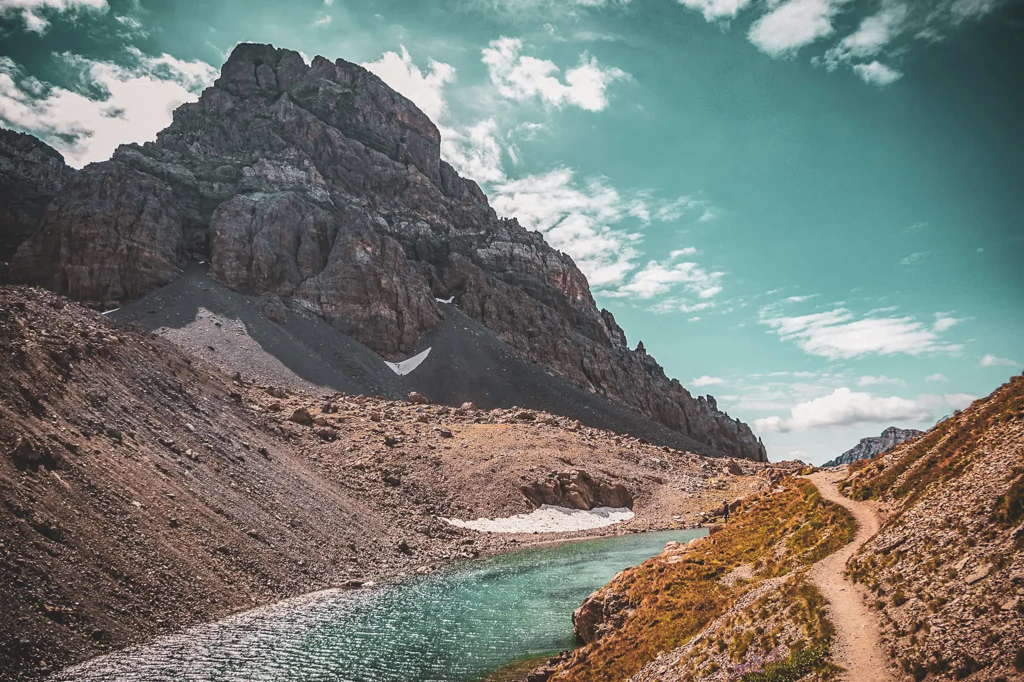 An impressive mountain landscape with craggy rocky peaks rising up under a blue sky dotted with clouds. A winding path cuts through an arid valley, bordered by the crystal-clear waters of a river.