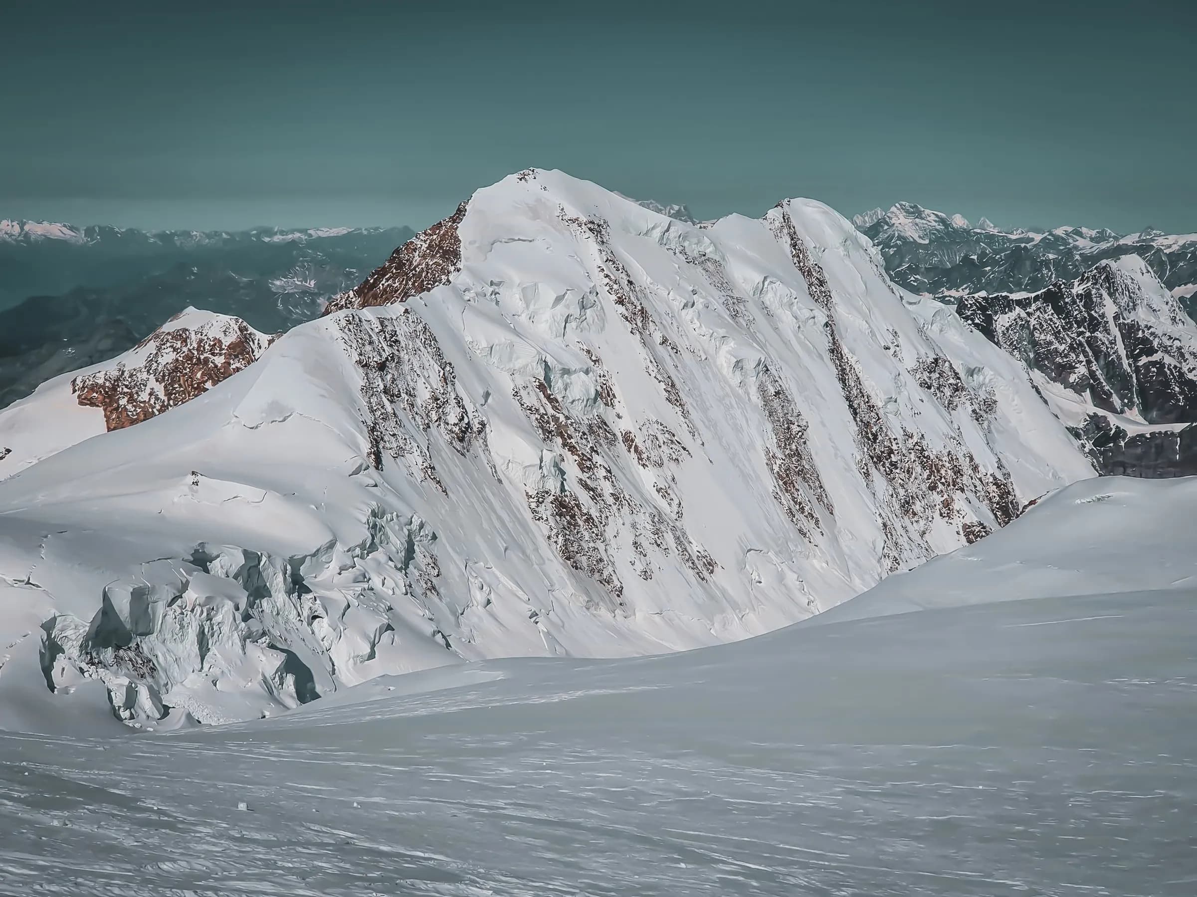 A landscape of snow-capped mountains, with majestic peaks covered in glistening snow. The peaks rise against a backdrop of blue-green sky, creating a striking contrast with the exposed rocks on some of the slopes.