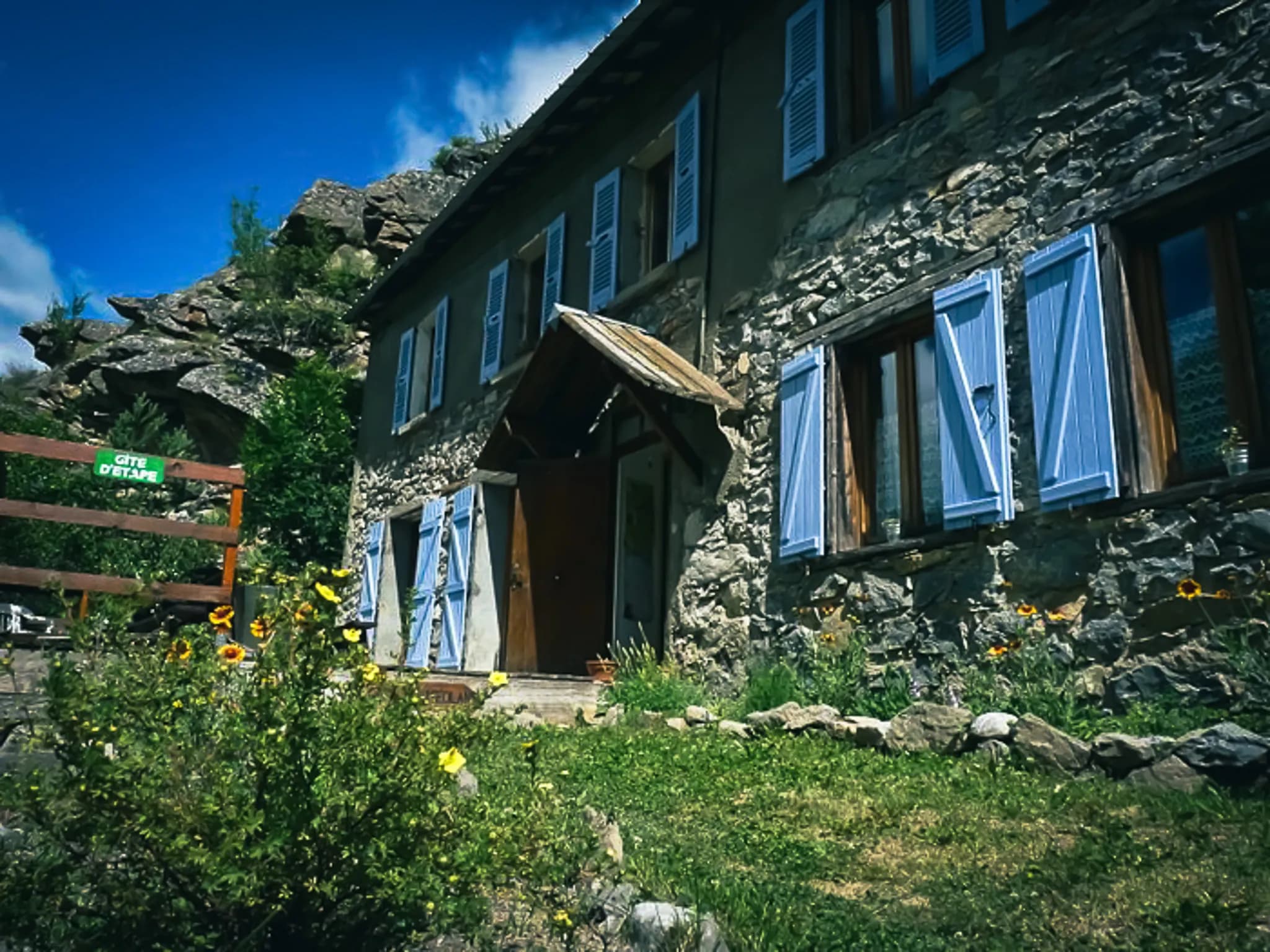 A stone mountain gîte with blue shutters, surrounded by greenery and colourful flowers. The entrance is welcoming, with a wooden awning, and a sign indicating "Gîte d'étape" is on the door.