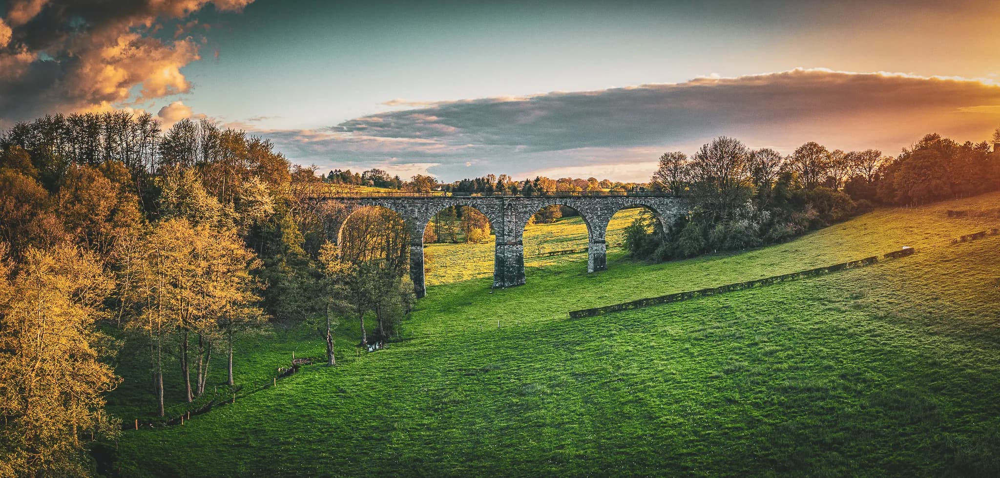 A stone viaduct overlooks a vast green field, surrounded by trees with golden leaves to mark the start of autumn. The sky is full of clouds, tinged with shades of orange and blue.