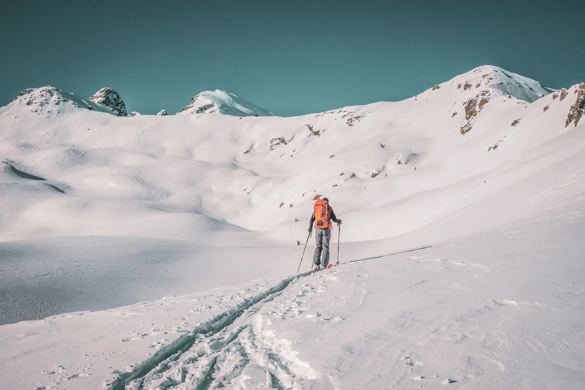 Un skieur évolue dans un paysage alpin recouvert d'une épaisse couche de neige, tandis que des montagnes majestueuses se dessinent en arrière-plan. Le skieur porte un grand sac à dos orange et utilise des bâtons