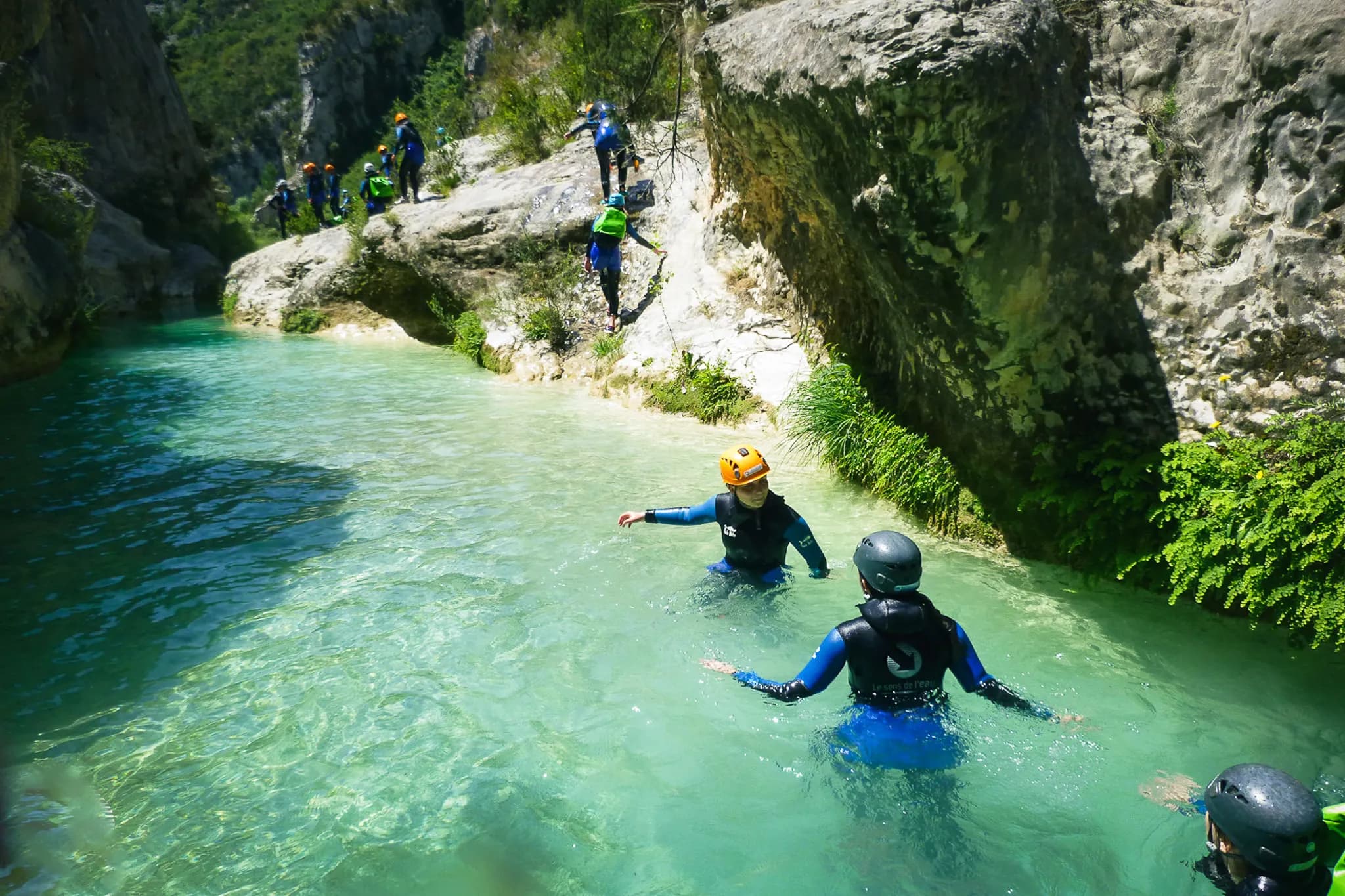 A canyoning scene in a narrow gorge with crystal-clear water. People in wetsuits, wearing orange and black helmets, move through the shallow water. A few participants climb