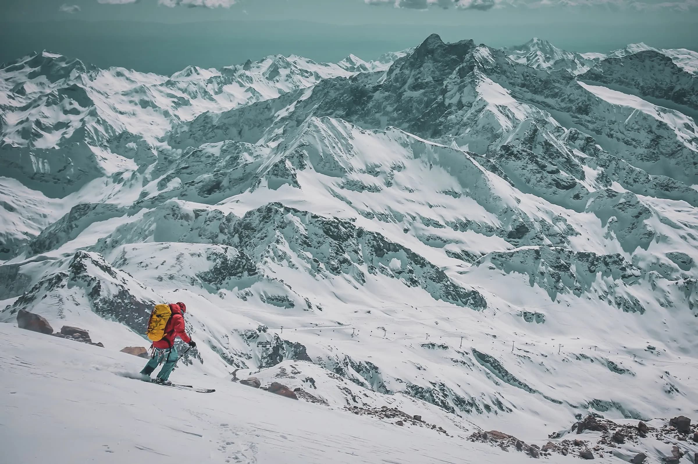 A skier in a red jacket and yellow rucksack descends a snowy slope, surrounded by majestic snow-capped mountains. Jagged peaks and deep valleys,