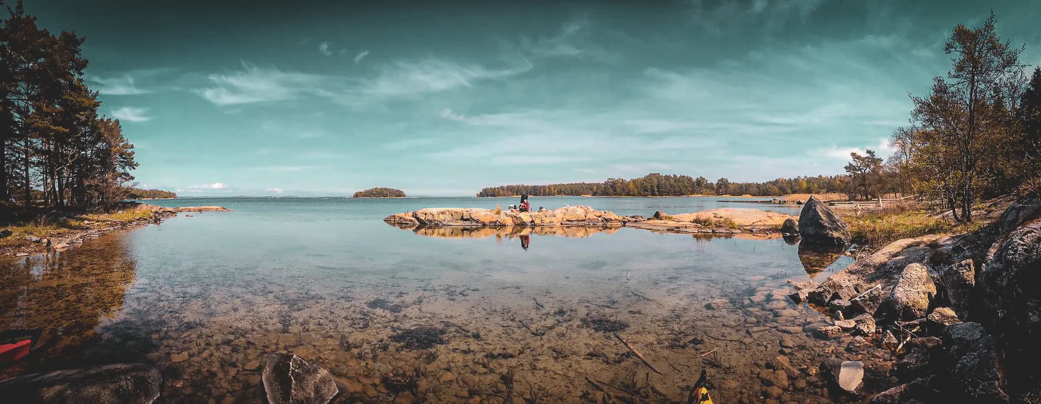 A serene landscape with calm waters reflecting the blue sky dotted with a few clouds. In the foreground, the water's edge is made up of rocks and stones, with areas of sand and a few grasses.