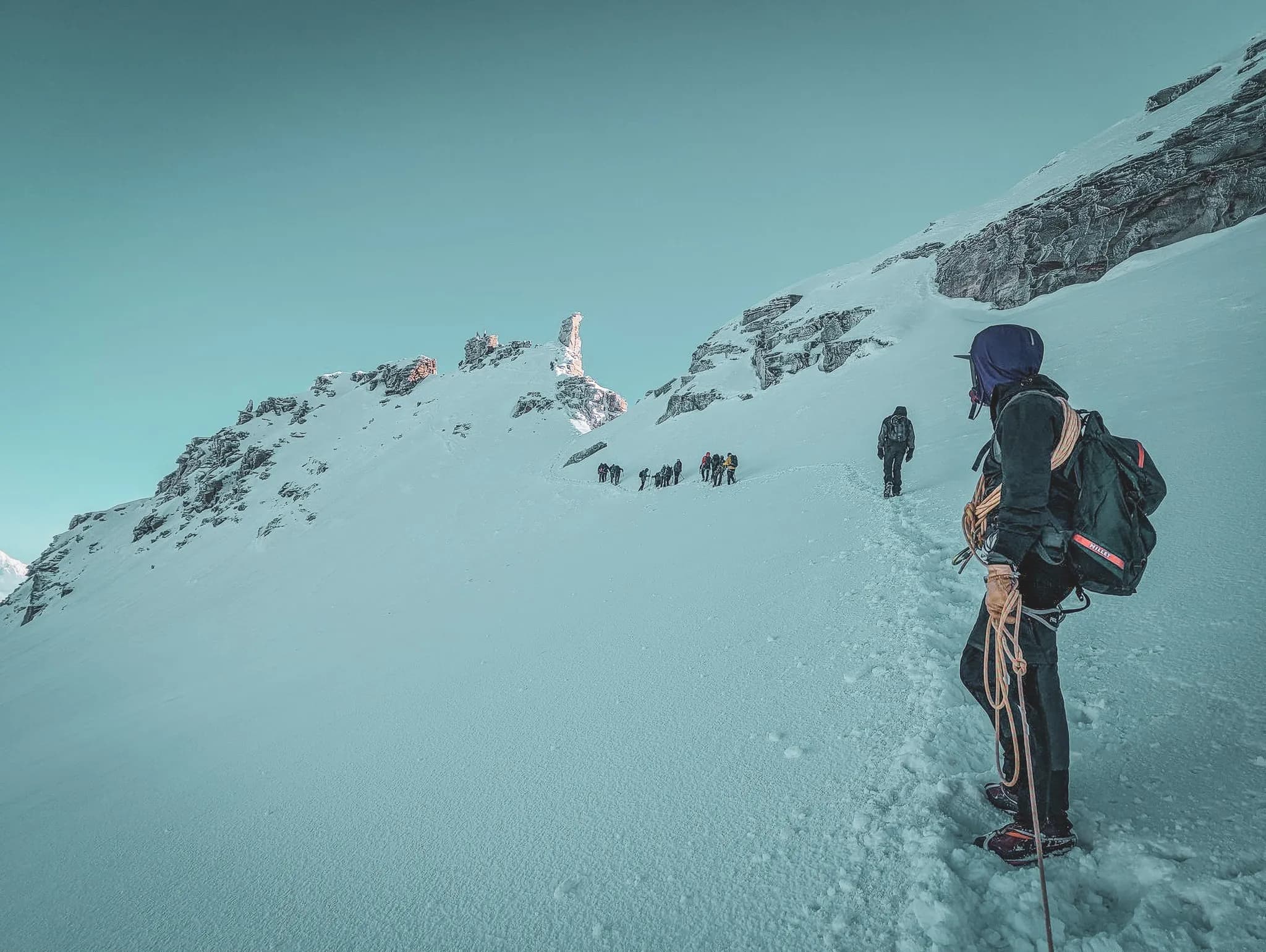 Een groep alpinisten beklimt de hellingen van Gran Paradiso, omringd door steile rotsen en een heldere blauwe lucht. Voorop draagt een klimmer een rugzak met touwen voor extra beveiliging.