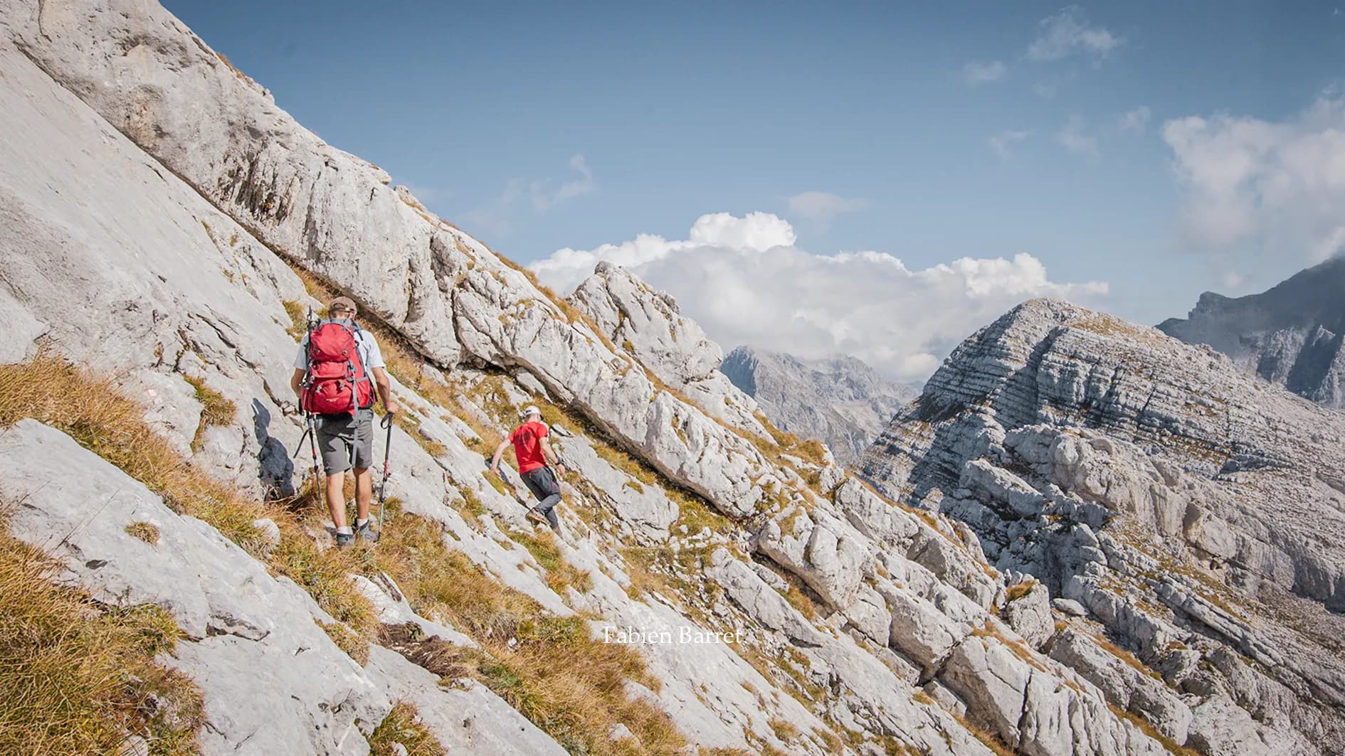 A mountainous landscape with rocky paths where two hikers are walking. On the left, a group with a large red rucksack is climbing up the side of a mountain, while on the right, a hiker in a t-shirt is walking along a rocky path.