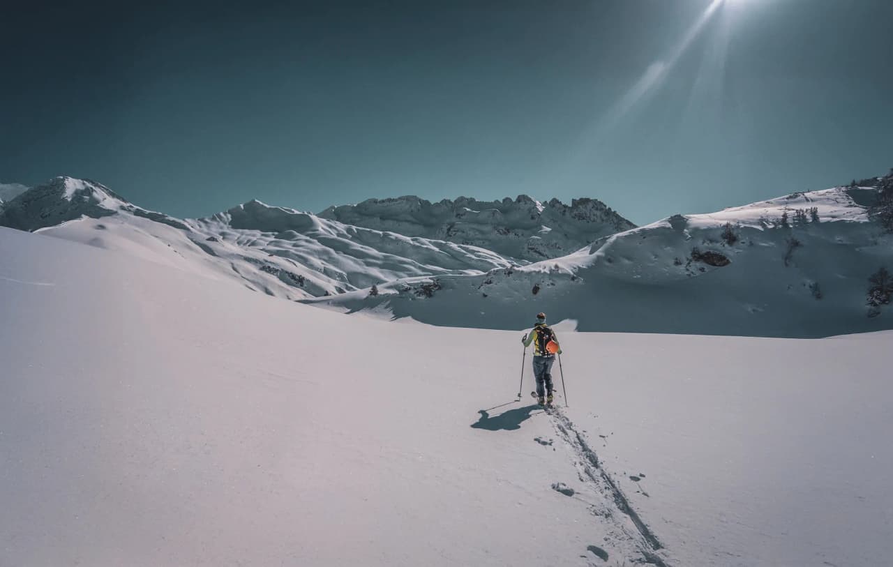 A lone skier moves through a vast snow-covered landscape, the tracks left by his skis marking the immaculate snow. In the background, majestic snow-covered peaks rise up to the horizon.
