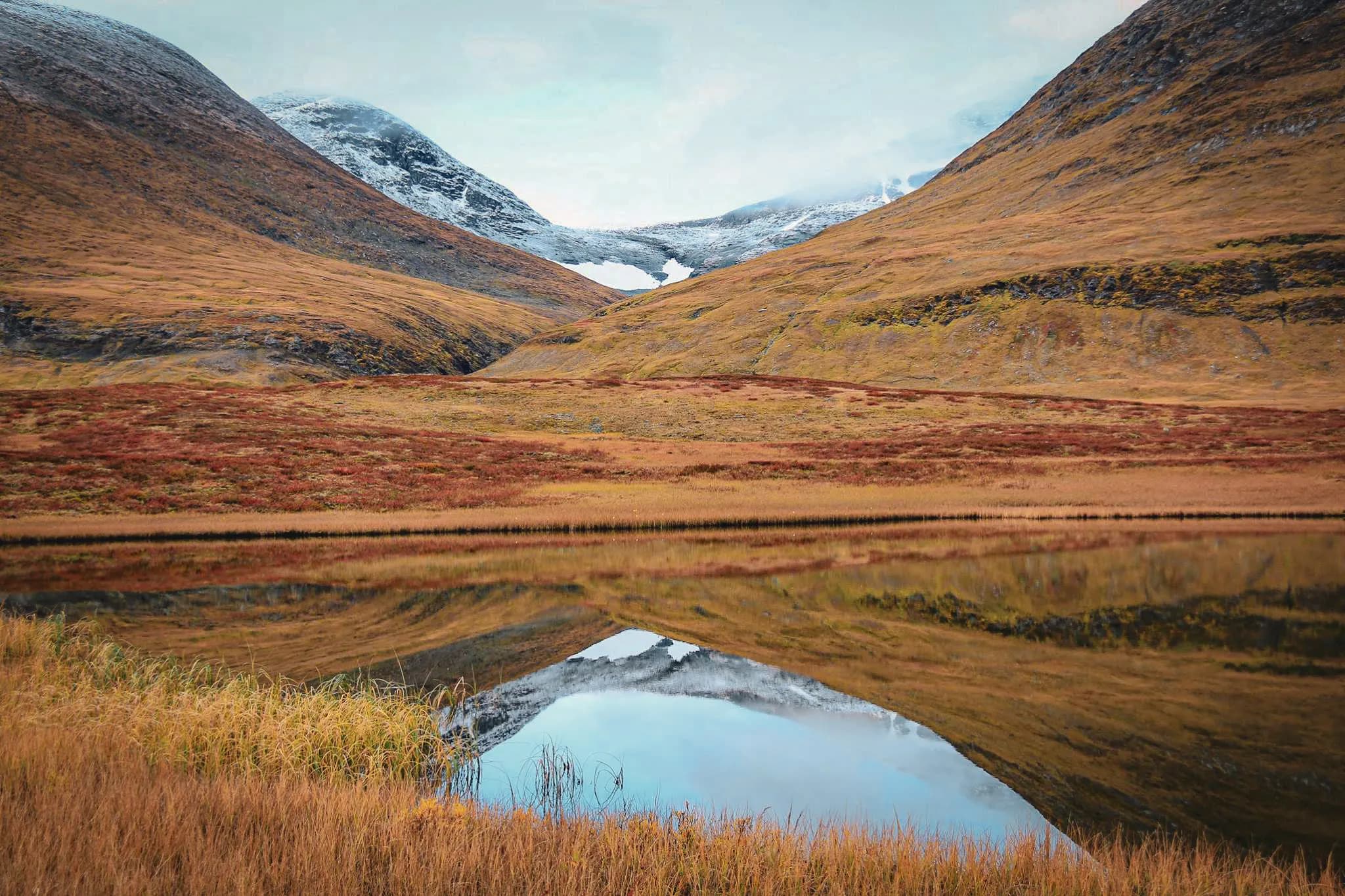 A mountain landscape of golden and brown hills, with snow-capped peaks in the distance. At the front, a calm lake reflects the mountains and cloudy sky, while