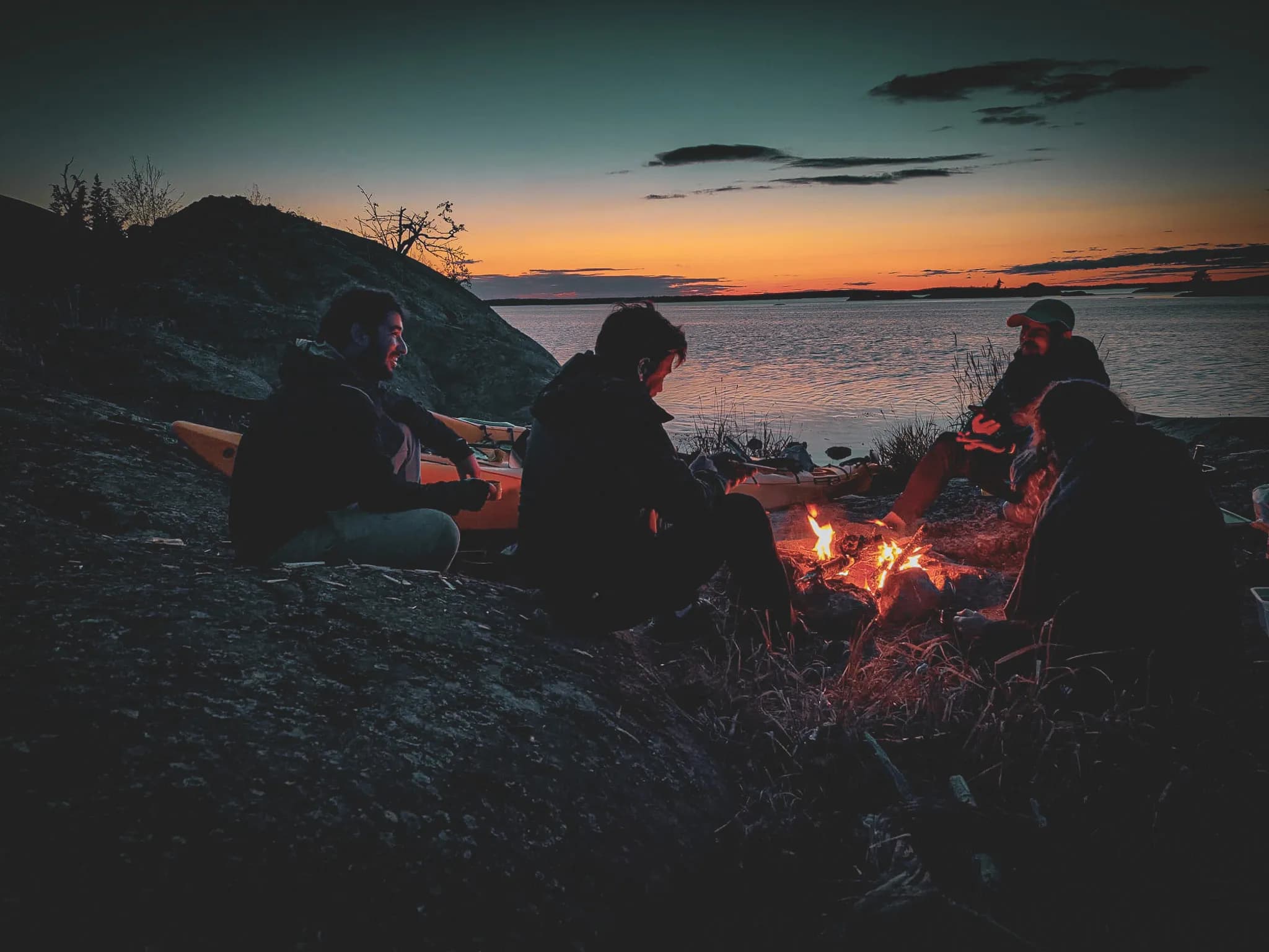 A group of people are sitting by a campfire on a riverbank, with a kayak visible nearby. The scene takes place at twilight, as the sun sets over the horizon, illuminating the sky with te