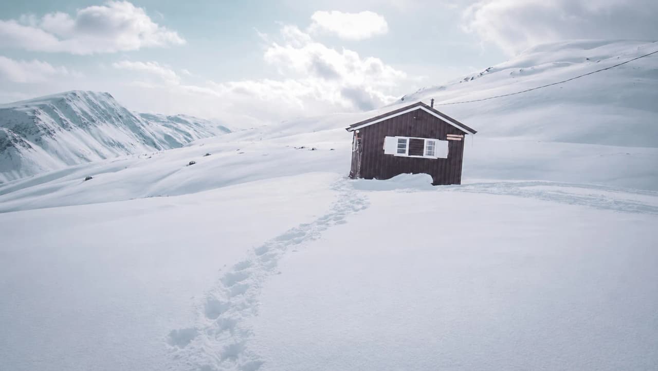 Een klein houten huisje, omringd door ongerepte sneeuw en bergen onder een blauwe hemel.