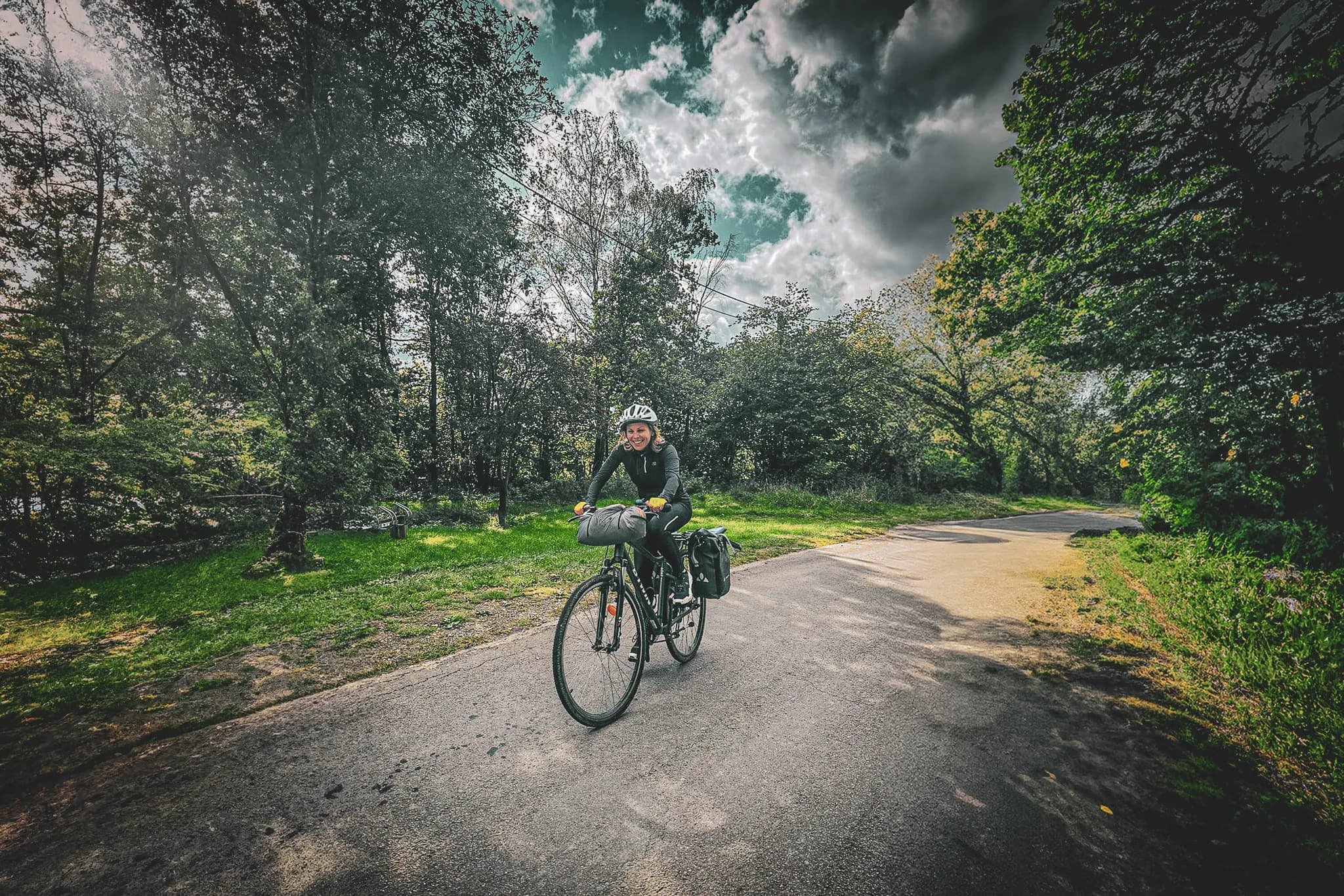 A smiling cyclist is riding along a cobbled road, surrounded by lush green vegetation and dense trees. He is wearing a safety helmet and appropriate cycling clothing. On his bike