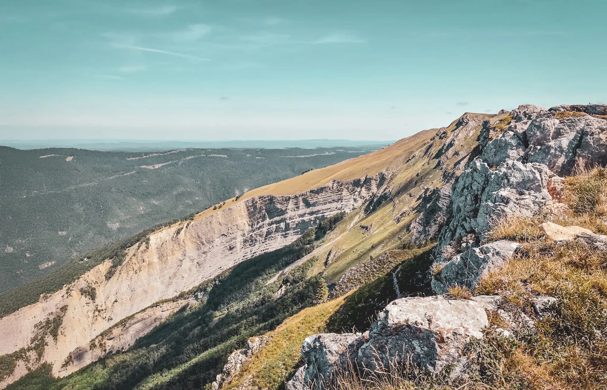 A mountainous landscape with steep cliffs, grassy slopes and rock formations. The view stretches across a wooded valley, and the sky is clear with a few light clouds. The contrast