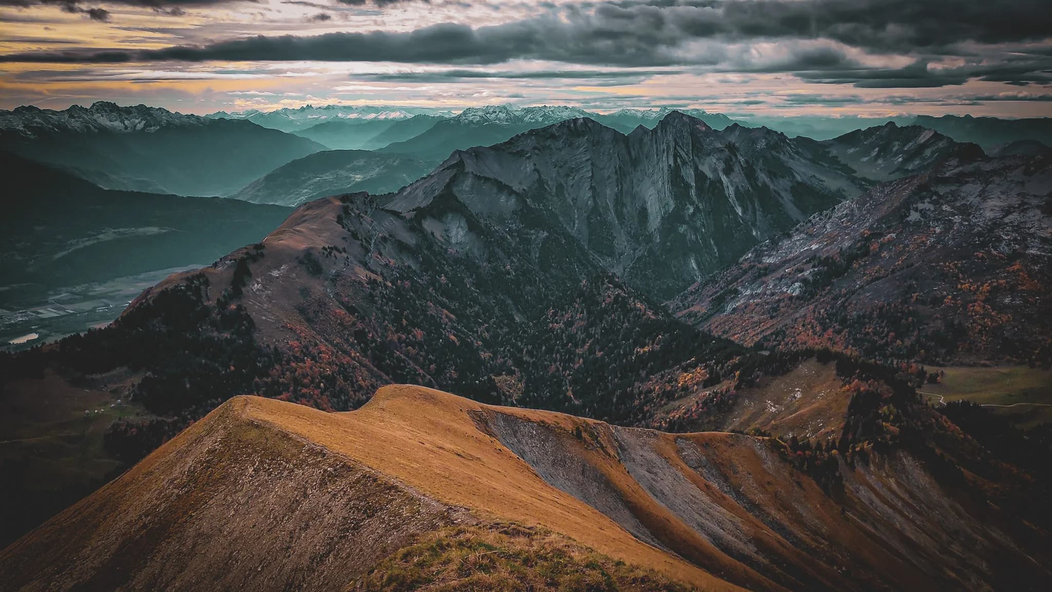 Un paysage montagneux impressionnant avec des sommets escarpés et des vallées verdoyantes. Les montagnes, partiellement enneigées, se dressent majestueusement sous un ciel nuageux. La lumière du soleil, filtrée