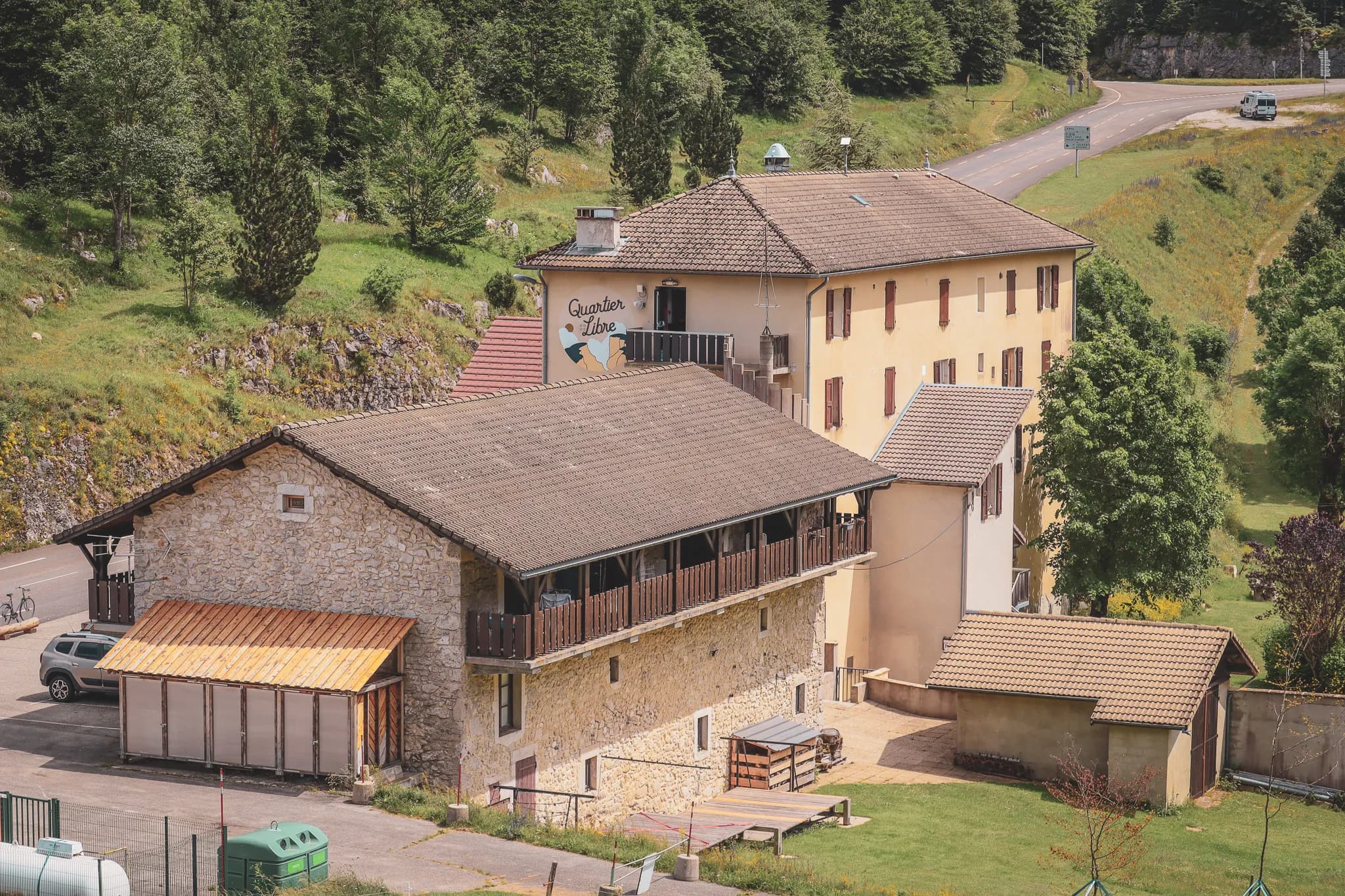 A building set in lush natural surroundings, with trees surrounding it. The architecture combines stone walls and painted facades, giving a rustic feel. The roof is tiled,
