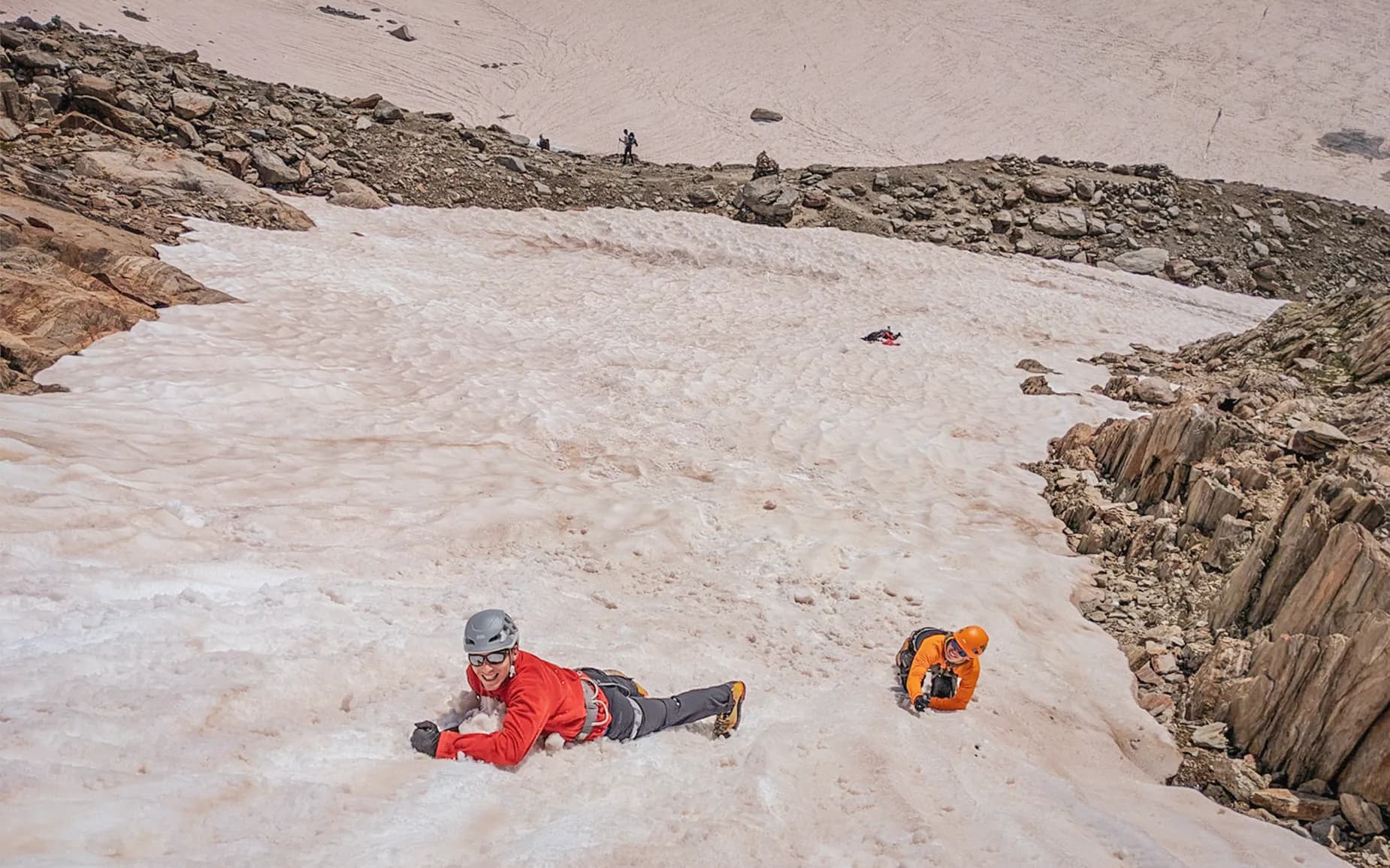 A glacier where people are climbing up a snowy slope. A climber at the top, wearing a red sweatshirt and a helmet, is smiling as he climbs onto the ice. Further down, another climber,