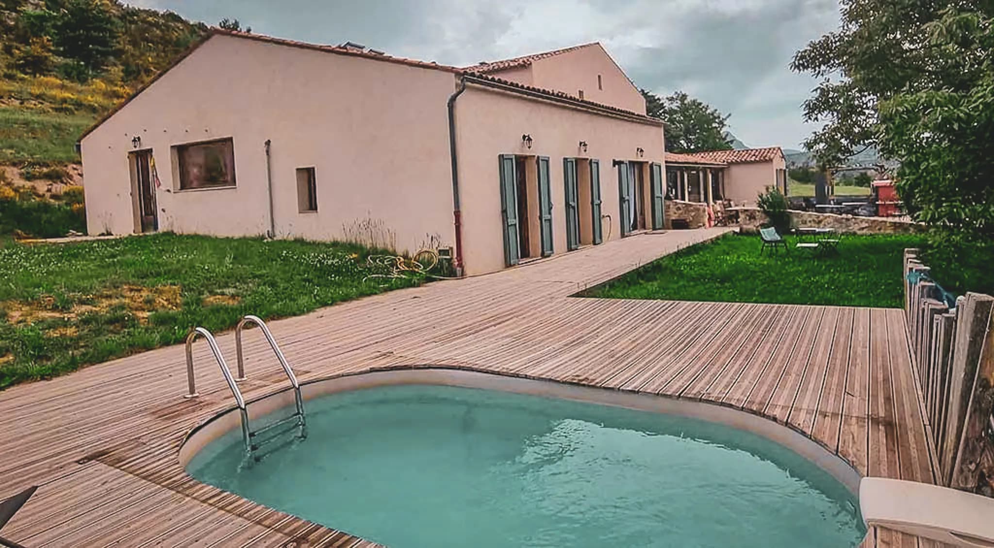 A modern house on the edge of a natural setting, surrounded by greenery. To the rear is a wooden outdoor area with a small swimming pool, overlooking a well-kept garden. The house has several windows with blue shutters