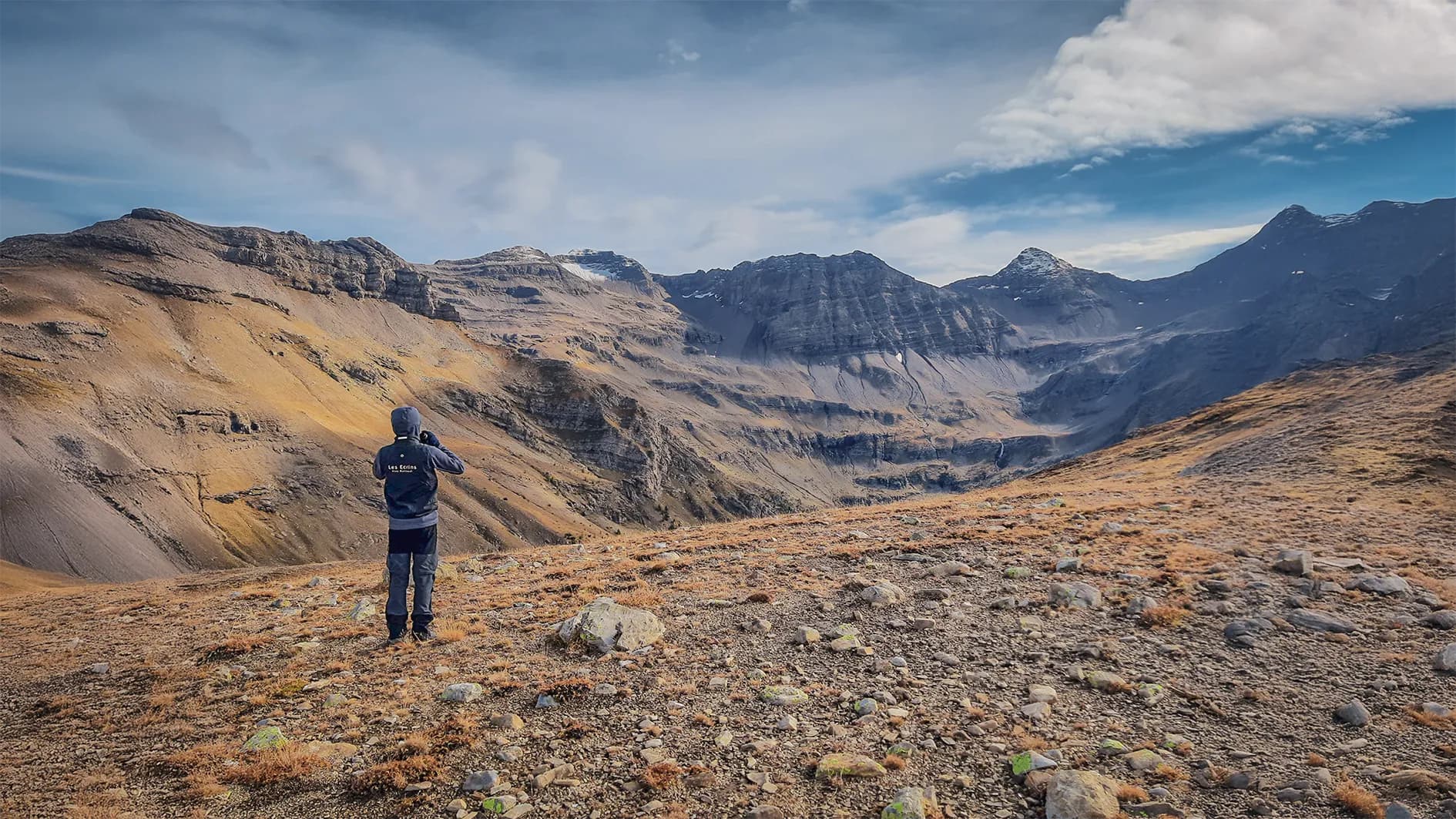 A man stands on rocky desert ground, looking out over an impressive mountain landscape. Sharply contoured mountains loom in the background, some covered in snow. The sky is part