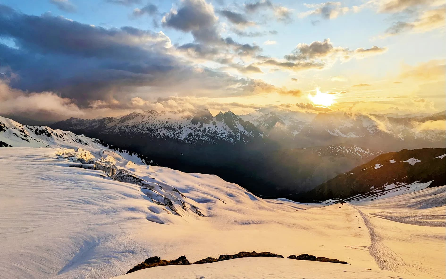 A spectacular mountain landscape at dusk, with snow-capped peaks beneath a dramatic sky dotted with clouds. The golden light of the setting sun shines down on the blanket of snow, creating shadows.