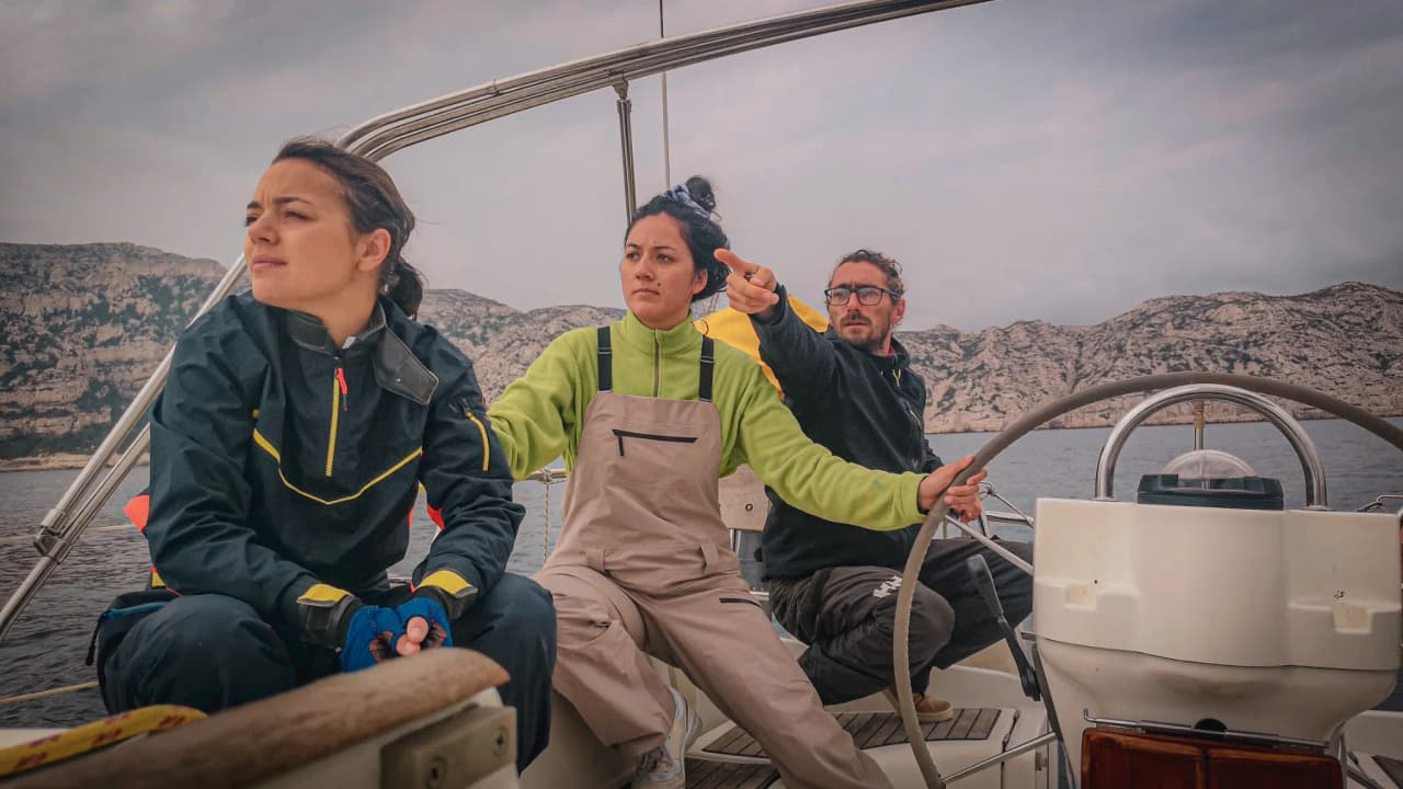 Drie mensen aan boord van een zeilboot, zeilend op de Middellandse Zee, met berglandschappen op de achtergrond.