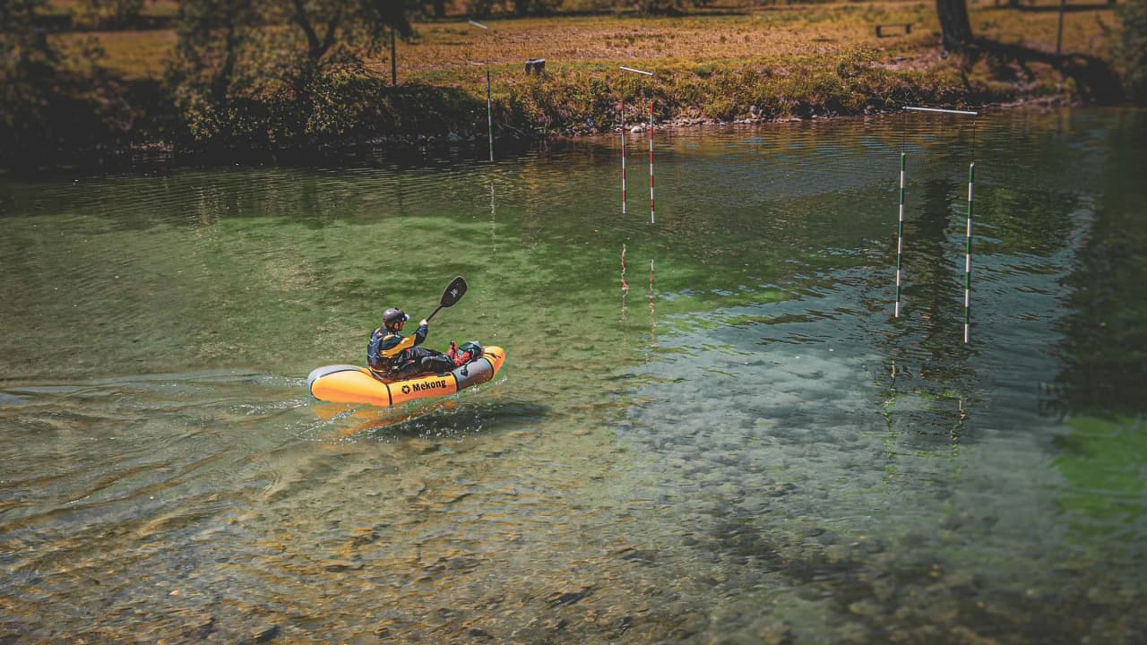 Een kanoër in een oranje packraft vaart over een kristalheldere rivier in de Alpen, omringd door groen.