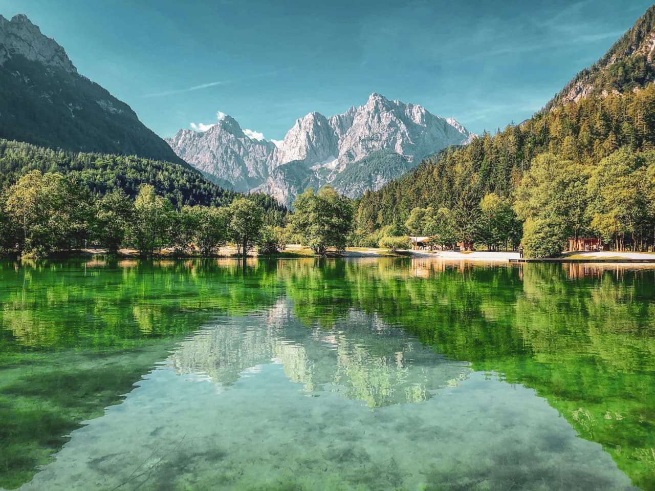 A tranquil lake surrounded by majestic mountains. The surface of the water reflects the mountain peaks and surrounding vegetation, creating a play of colours ranging from emerald green to light blue. Lush trees