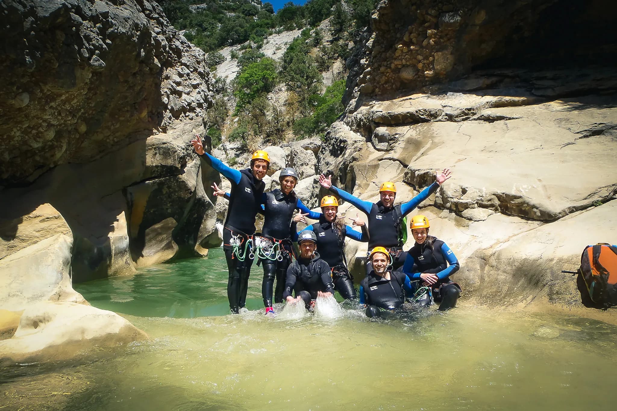 A group of smiling people stand in a small clearing near the water, surrounded by large rock formations. They are wearing black wetsuits and yellow safety helmets. Some members of the group