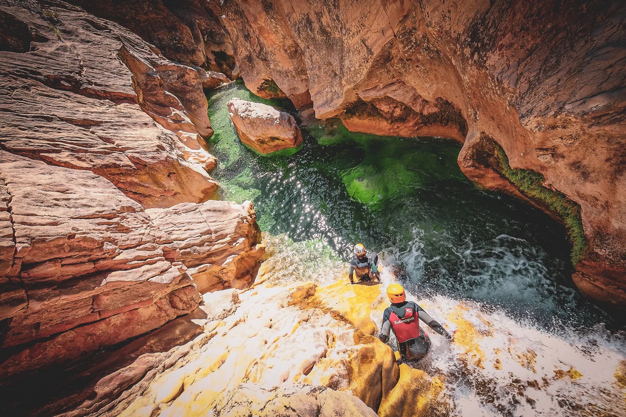 A narrow canyon with rocky walls, large granite formations and ochre hues. Two people in wetsuits swim beside a stream of water whose reflections are a dazzling green.
