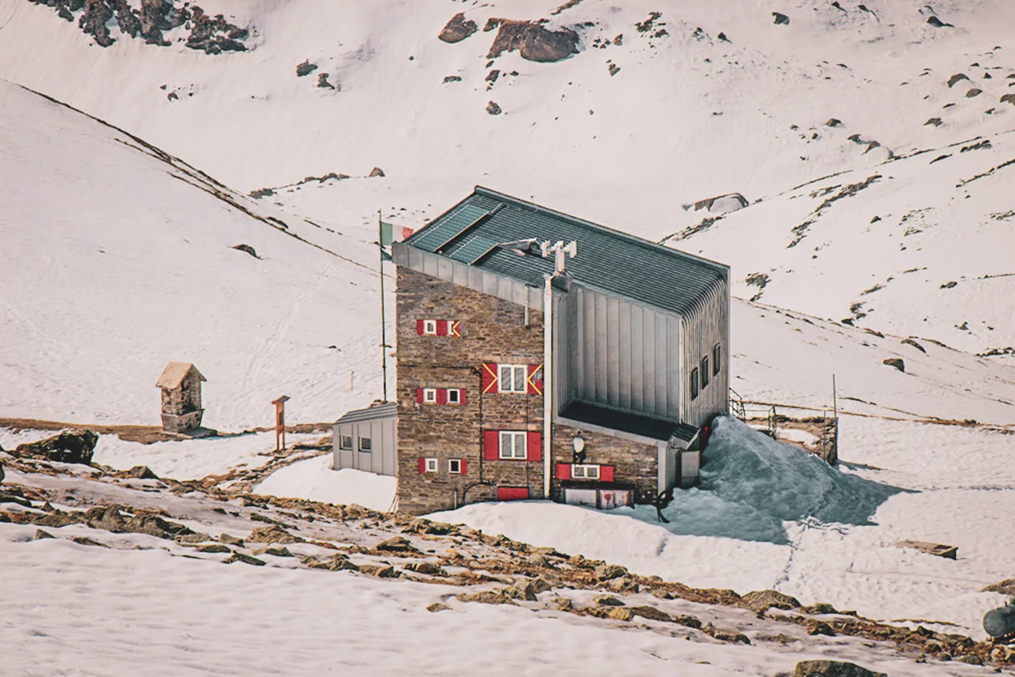 A modern building stands on snow-covered ground, surrounded by mountains. The building has a stone and metal structure, with brightly coloured windows and decorations. Nearby, a small wooden hut