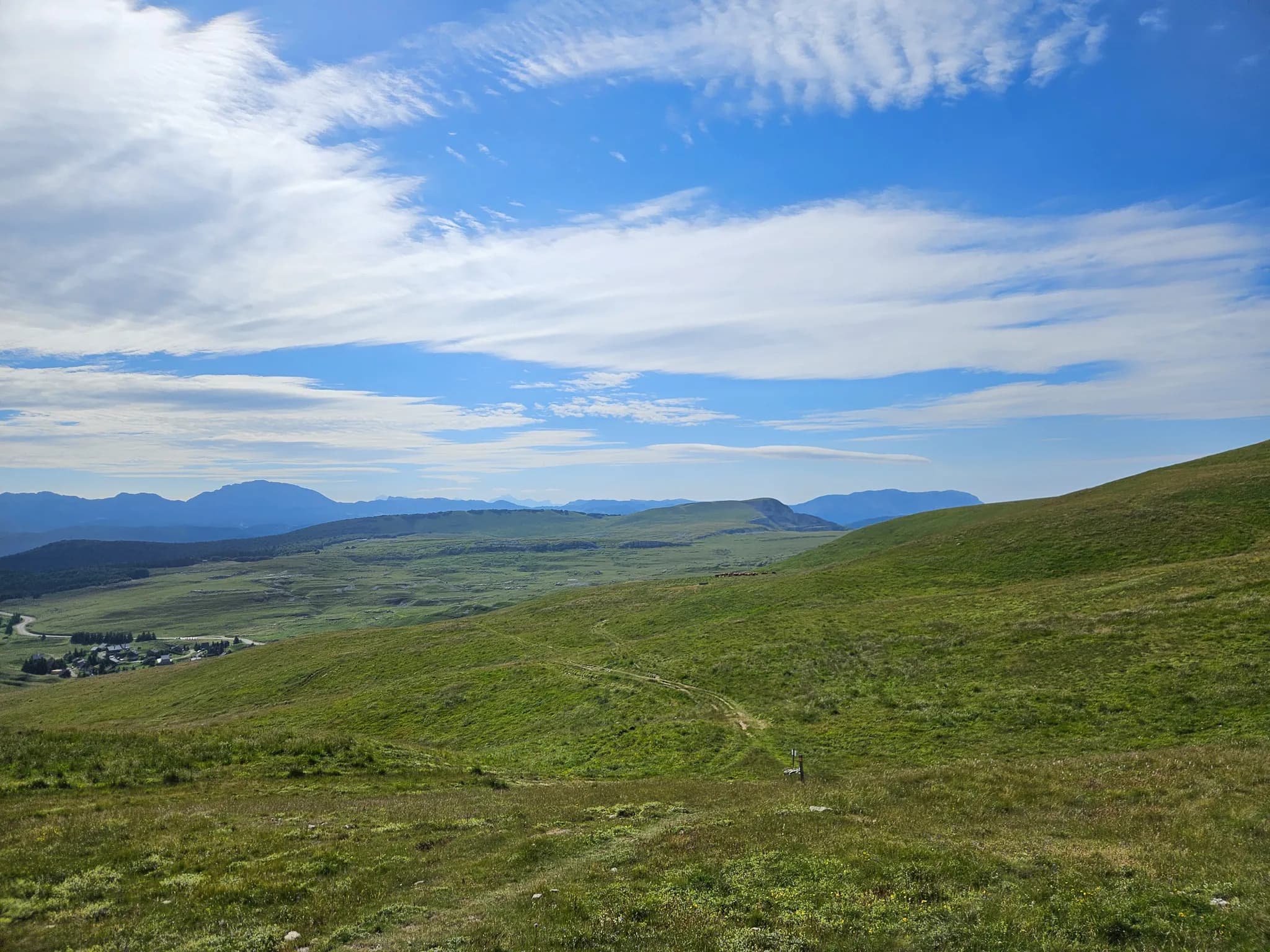 A mountainous landscape with green hills stretching to infinity. The sky is clear with scattered white clouds, creating a pleasant contrast with the green of the meadows. In the background