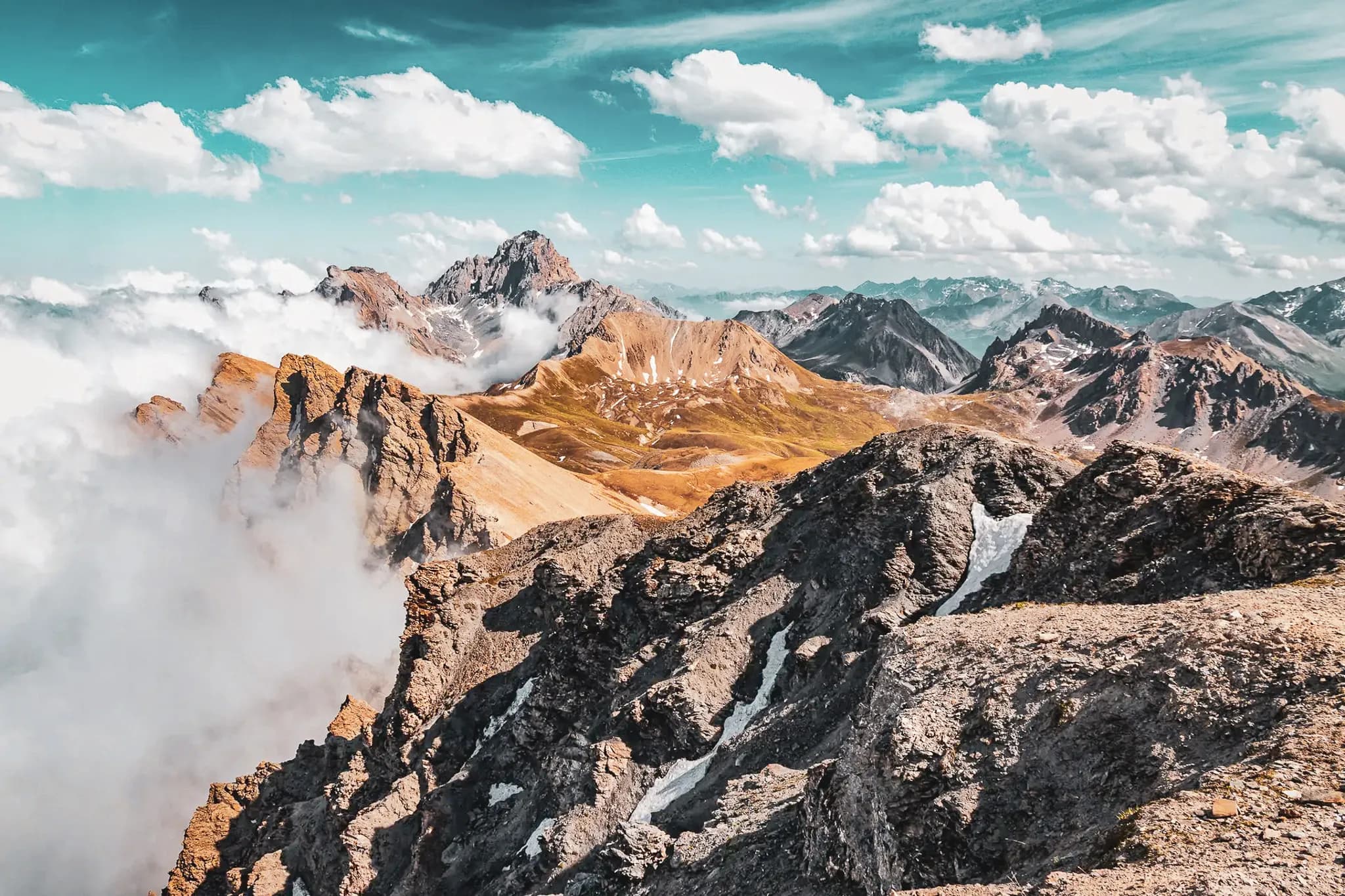 A spectacular mountain landscape under a blue sky dotted with white clouds. Steep peaks rise majestically, surrounded by light mist and golden valleys. The rocky slopes of