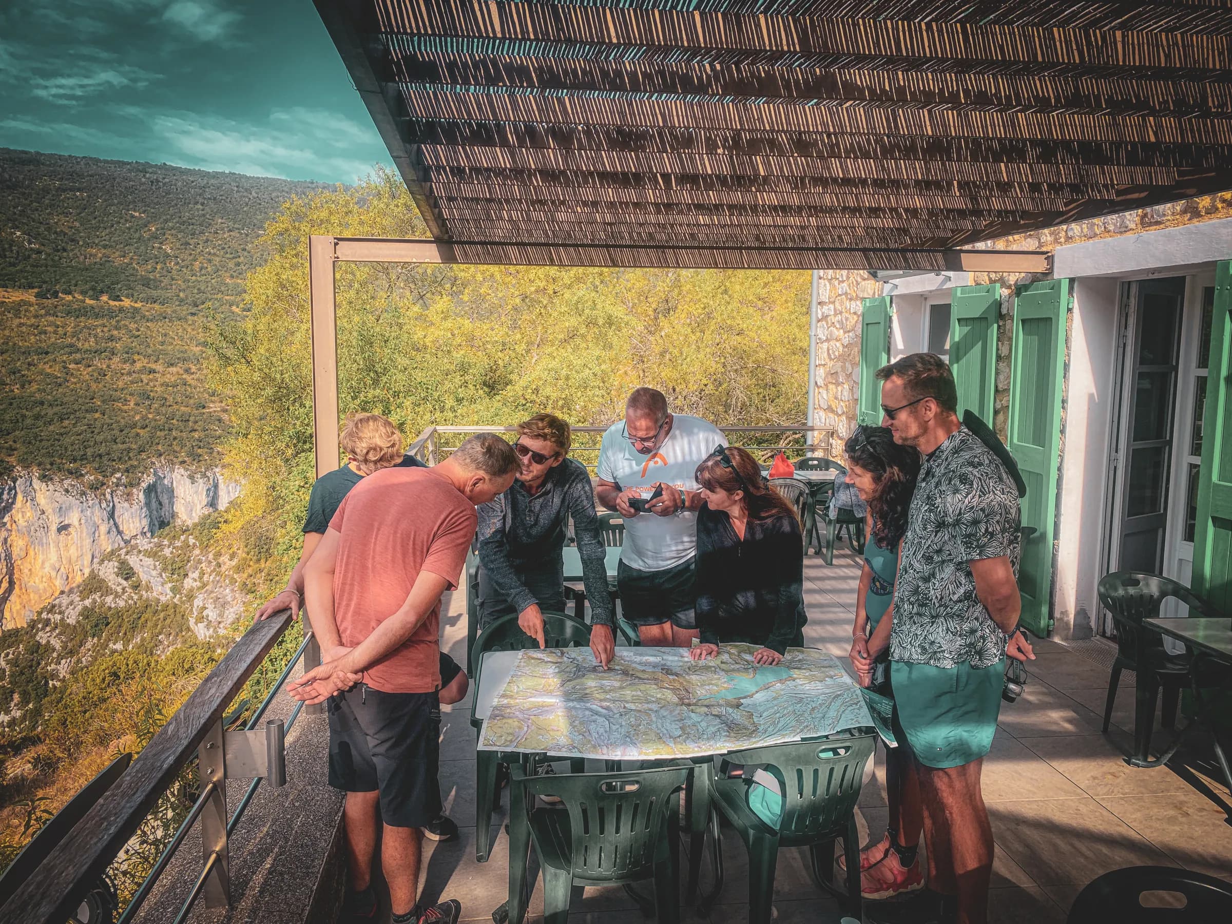 A group of people are looking at a large map placed on a table on a terrace. The setting is mountainous, with green hills in the background. The participants, dressed casually, appear to be discussing