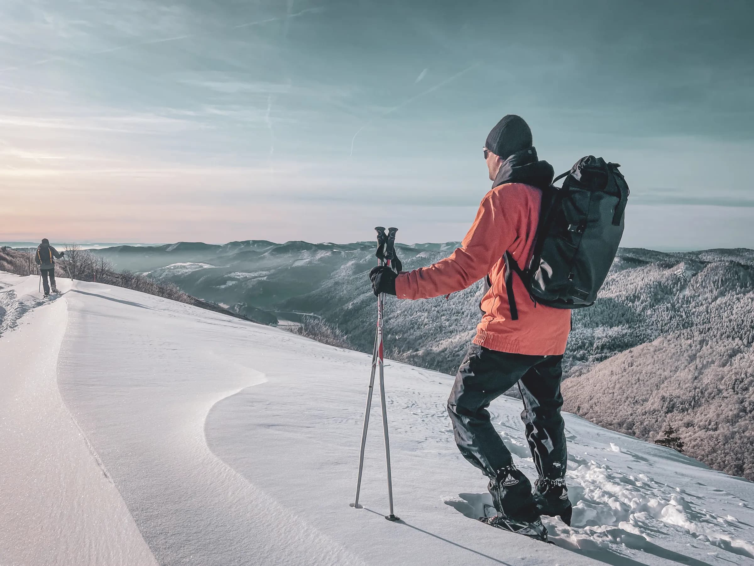 A hiker on the snow, wearing an orange jacket and black trousers, is standing on a summit. He is carrying a large rucksack and snowshoes, holding hiking poles. In the background, a