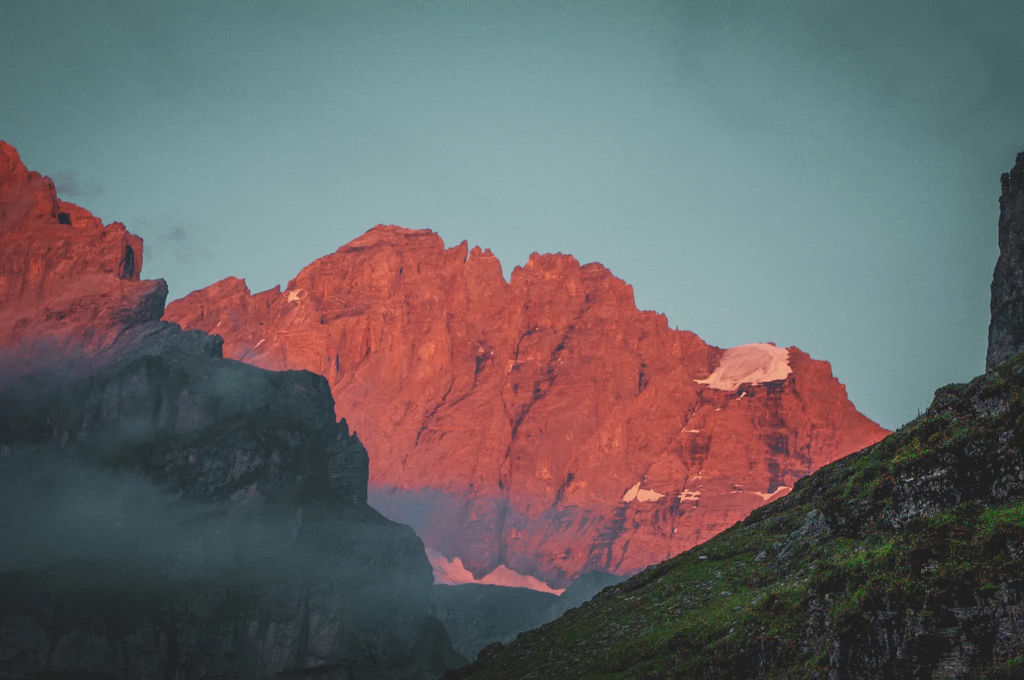 A mountainous landscape at dusk, with steep peaks illuminated by red and orange hues, creating a striking contrast with the clear blue sky. Subtle clouds envelop the bases of the mountains.