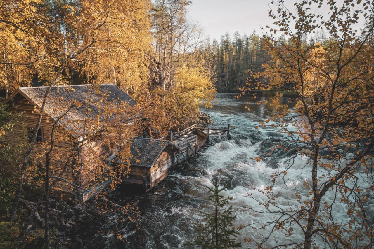 A wooden hut on the banks of a tumultuous river, surrounded by trees with golden leaves.