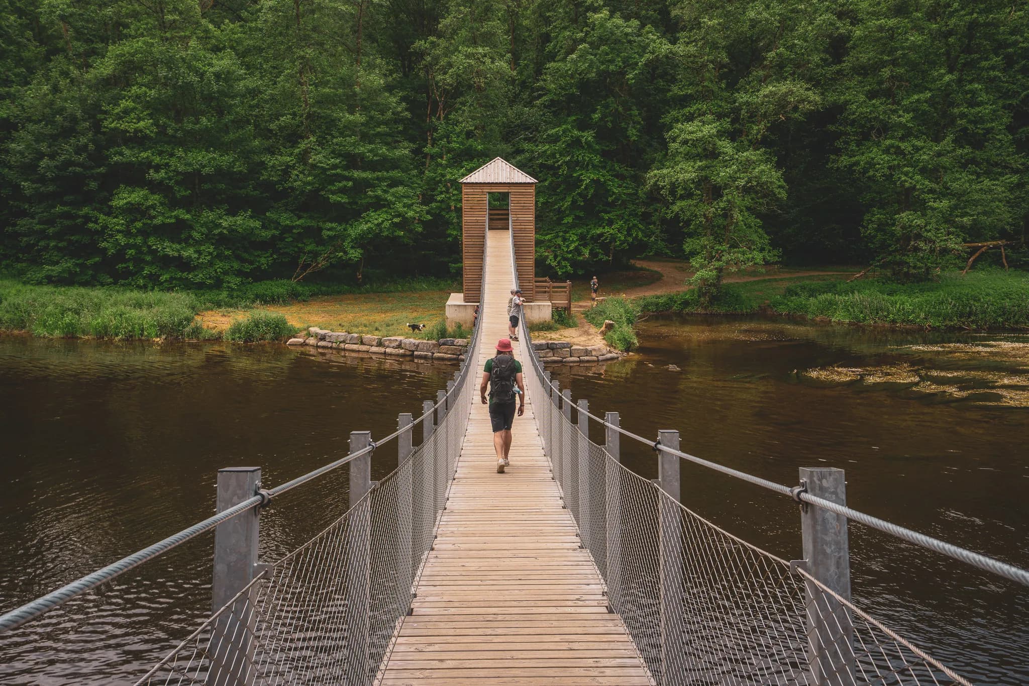 Een houten hangbrug overspant een kalme rivier, omgeven door een dicht groen bos. In de verte is een kleine houten hut te zien op de top van een heuvel, bereikbaar via de brug. Mensen wandelen