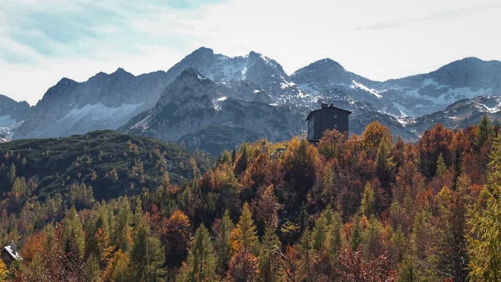 A black chalet stands at the top of a hill, surrounded by a forest of autumnal colours. The trees are shades of red, orange and yellow, creating a vibrant contrast with the green of the con