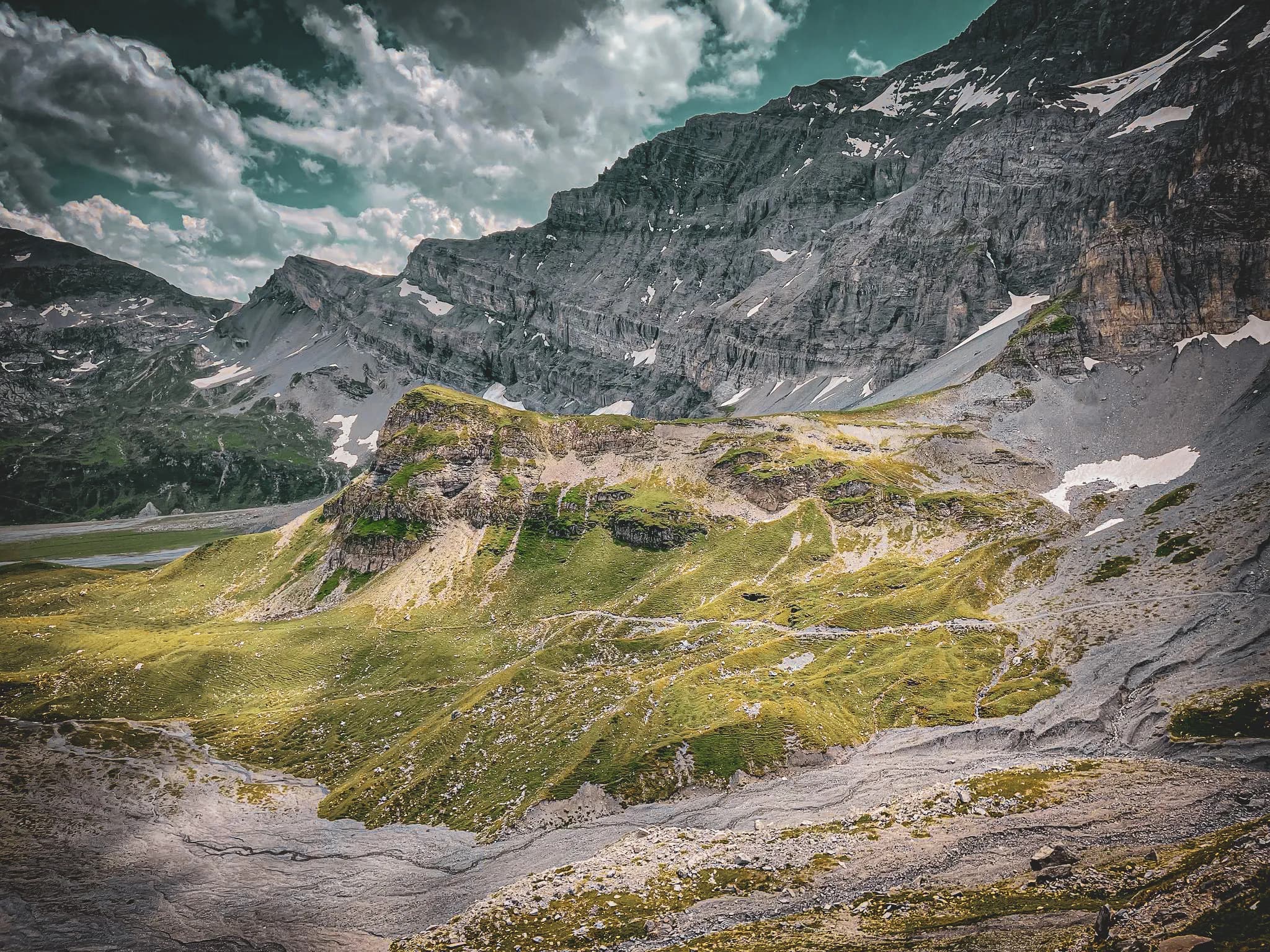 A mountainous landscape characterised by steep slopes and bare rock. The vegetation consists mainly of green areas, with traces of snow on the summits. The sky is dotted with