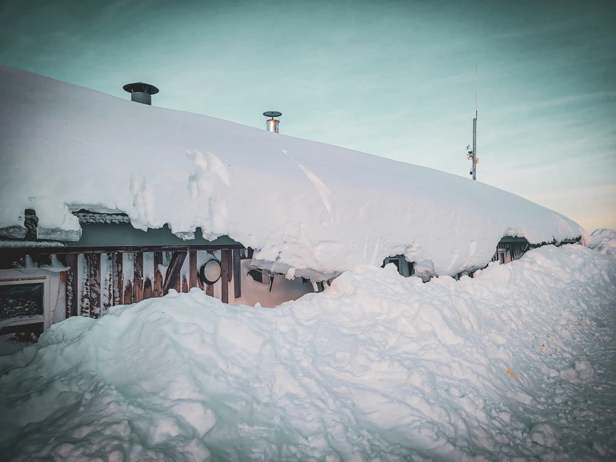 Een gebouw bedekt met sneeuw, met een schuin dak bedekt met een dikke laag sneeuw. Schoorsteenpijpen komen tevoorschijn uit de sneeuw, terwijl hoopjes sneeuw kleine hoopjes vormen rond het gebouw.