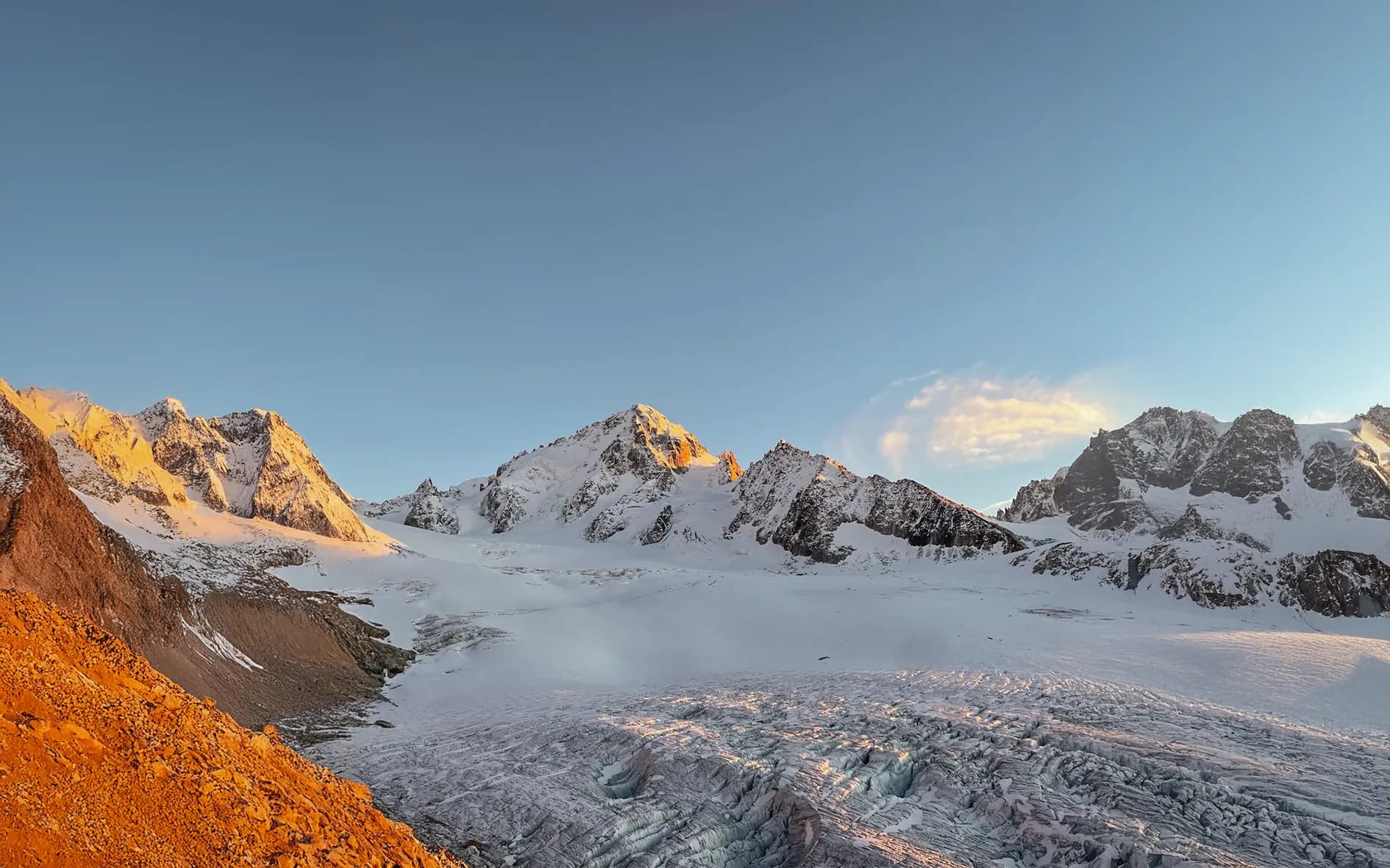 A majestic Alpine landscape with snow-capped peaks illuminated by the morning light. In the foreground, a vast, winding glacier covers the ground, while the surrounding rocks are shades of orange.